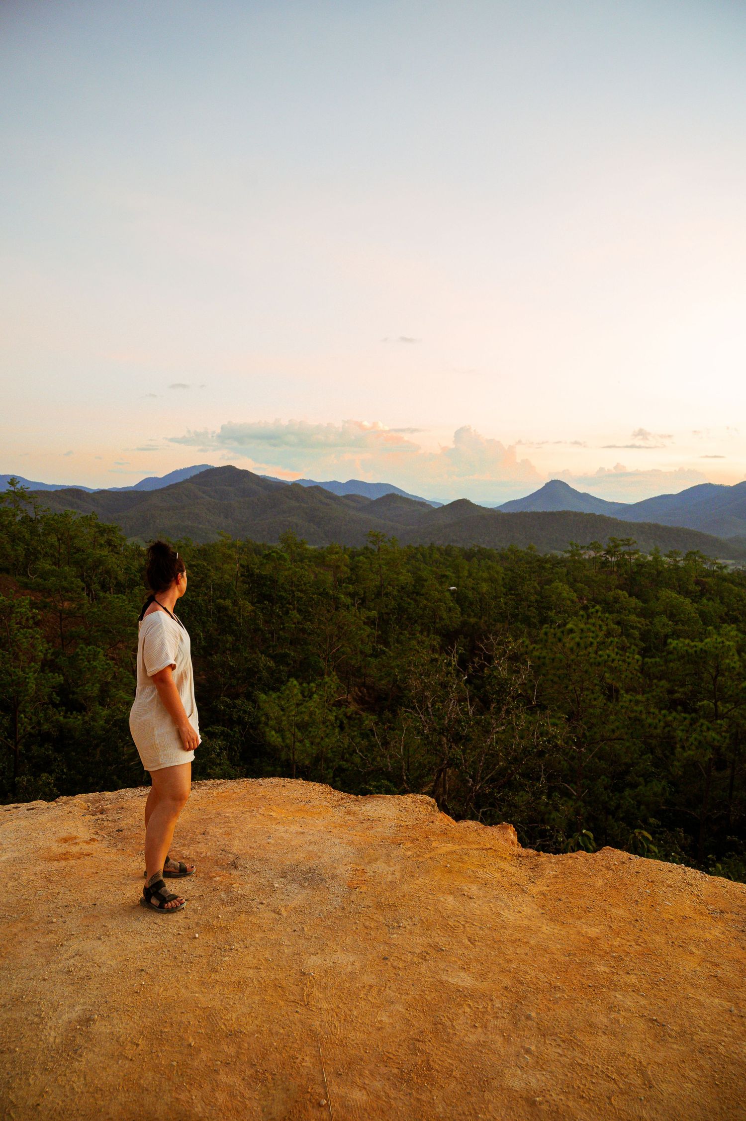 A person stands on a rocky overlook gazing at mountain ranges and dense forest during golden hour.