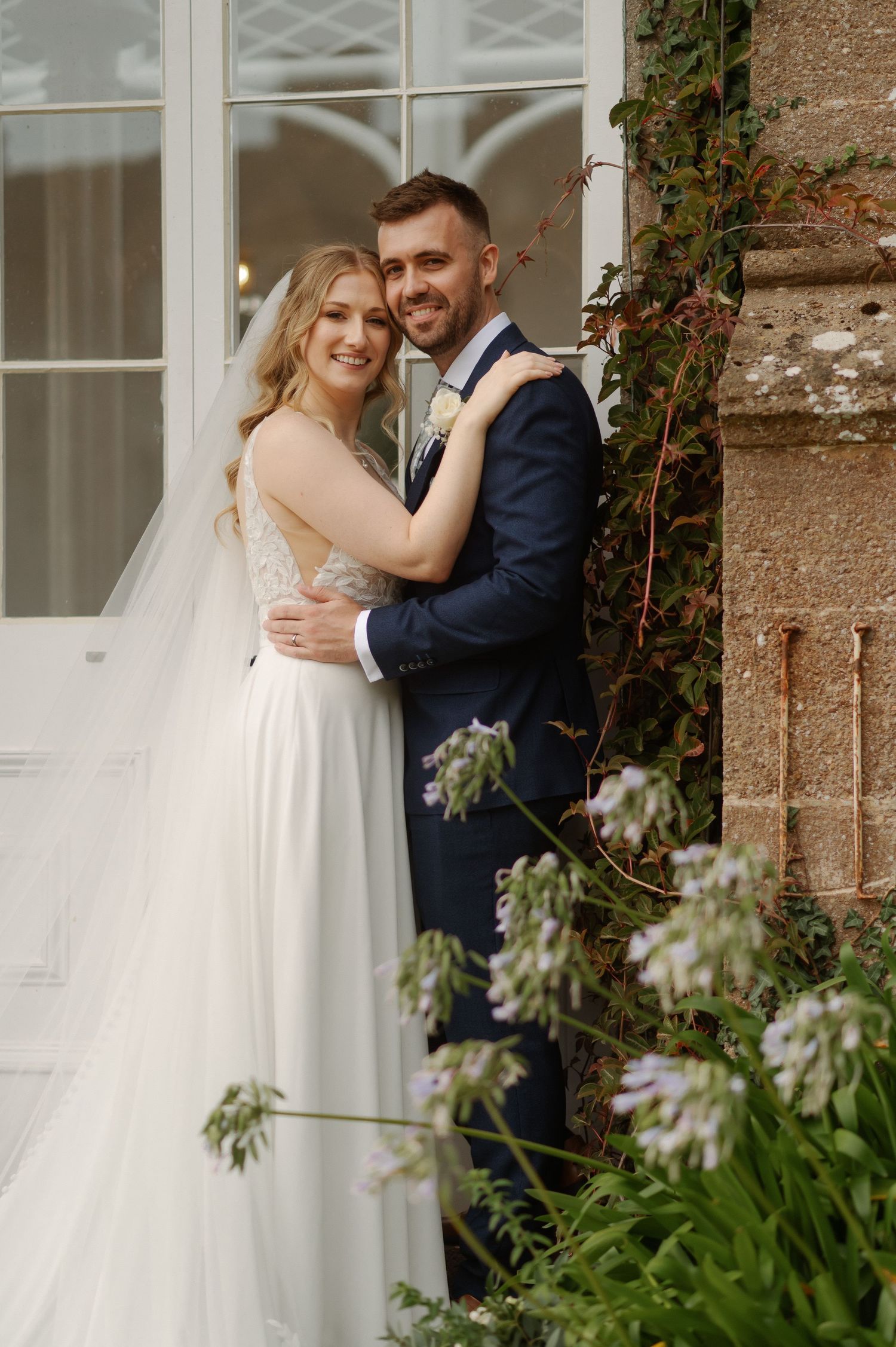 A romantic wedding couple embraces by a stone wall with climbing vines and white flowers in the garden.