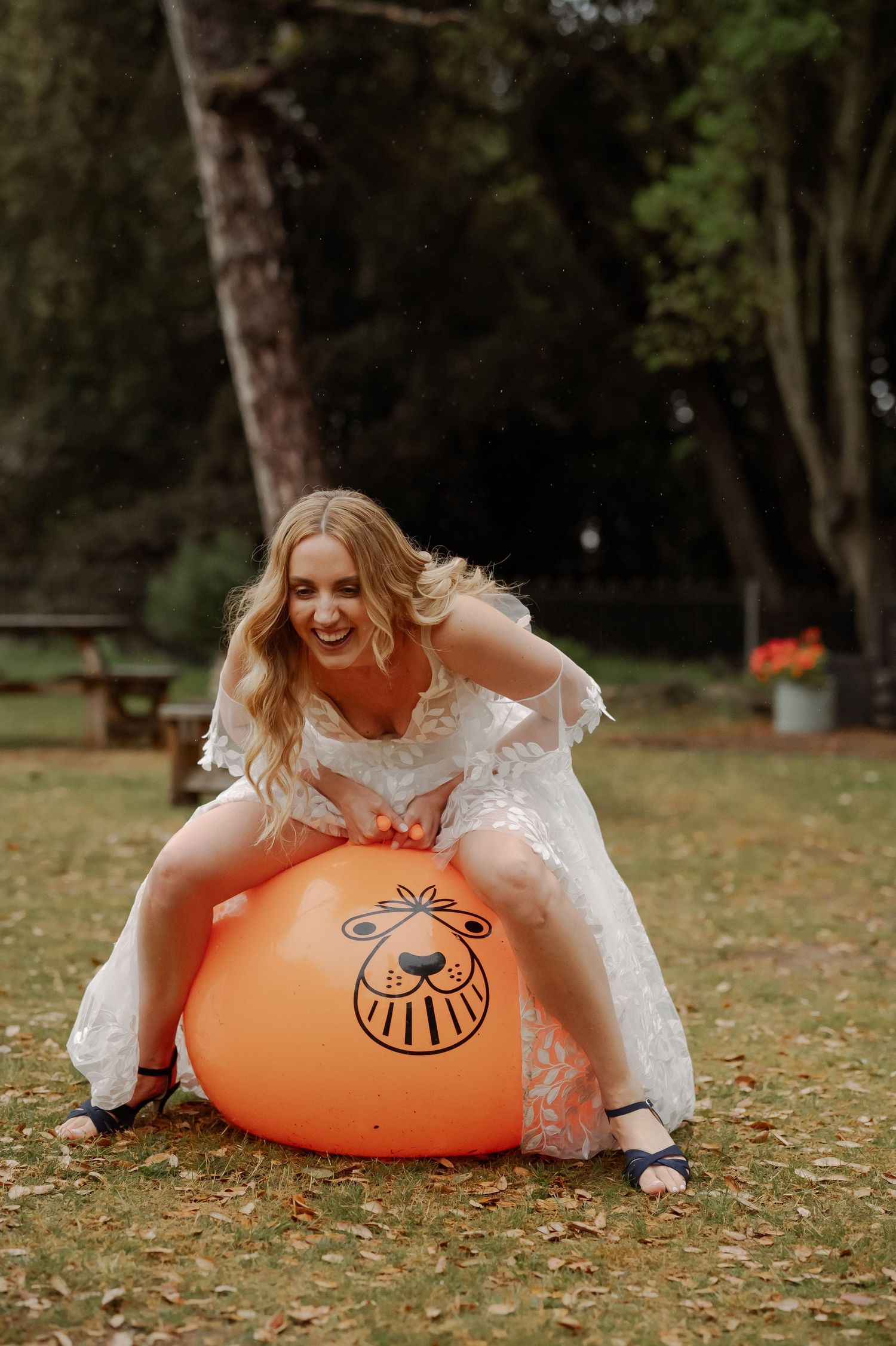 A person in a white dress playfully bounces on an orange space hopper toy in an autumn park setting.