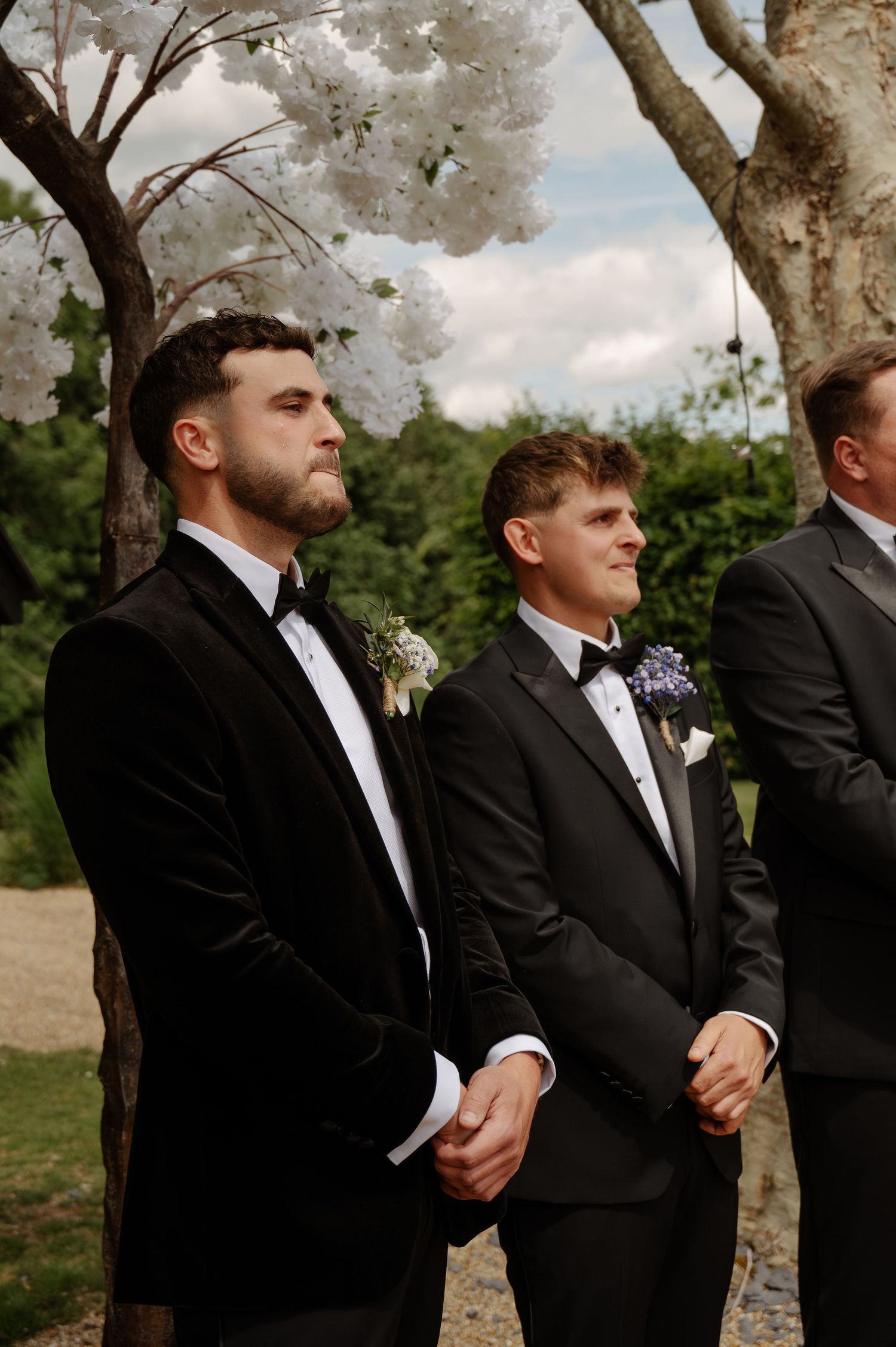 Groomsmen in black tuxedos stand together outdoors during a formal wedding ceremony.