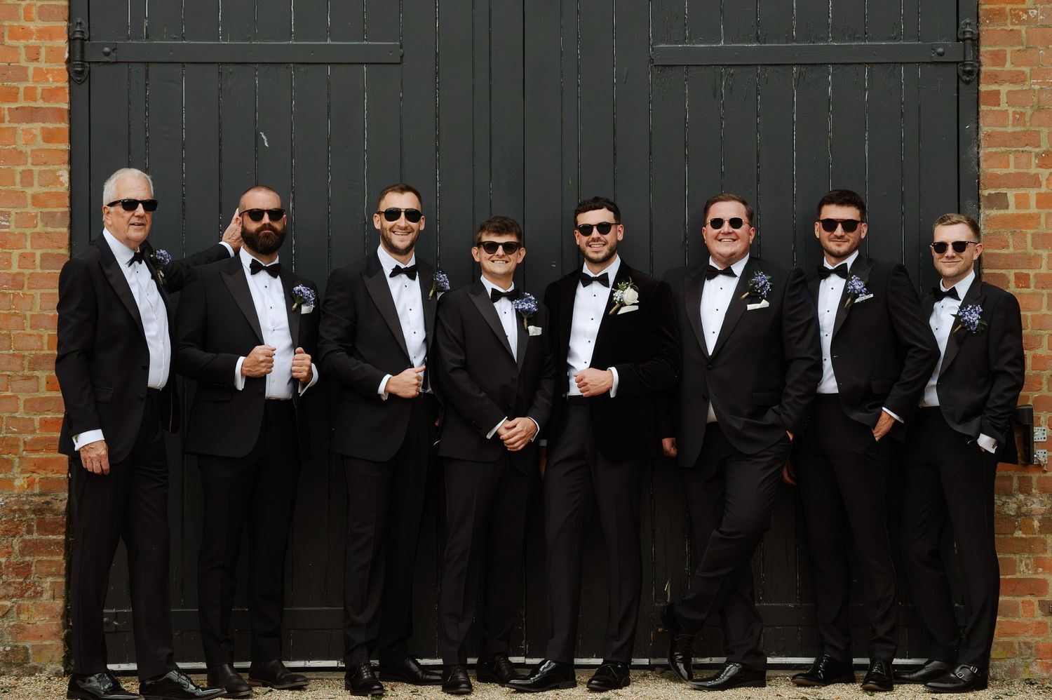Wedding party in black tuxedos and sunglasses pose against brick wall with dark doors.