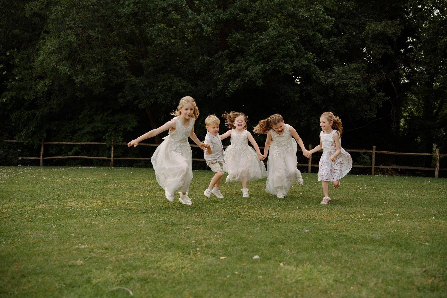 Wedding party in white dresses running playfully across a lush green lawn during a summer celebration.