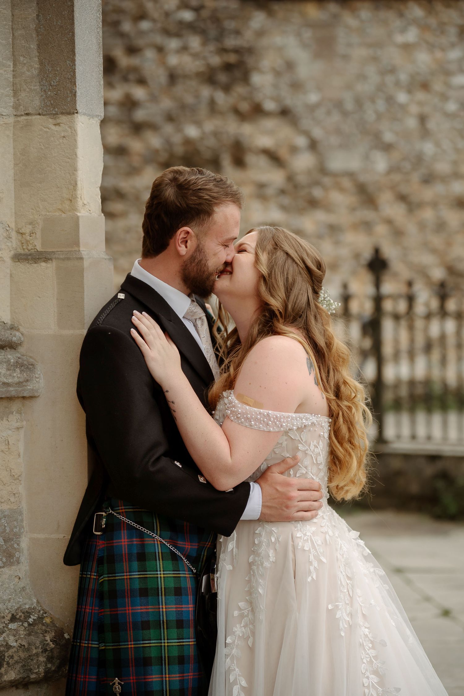 Bride and groom sharing a romantic kiss, groom wearing a tartan kilt, bride in an off-shoulder lace wedding gown.