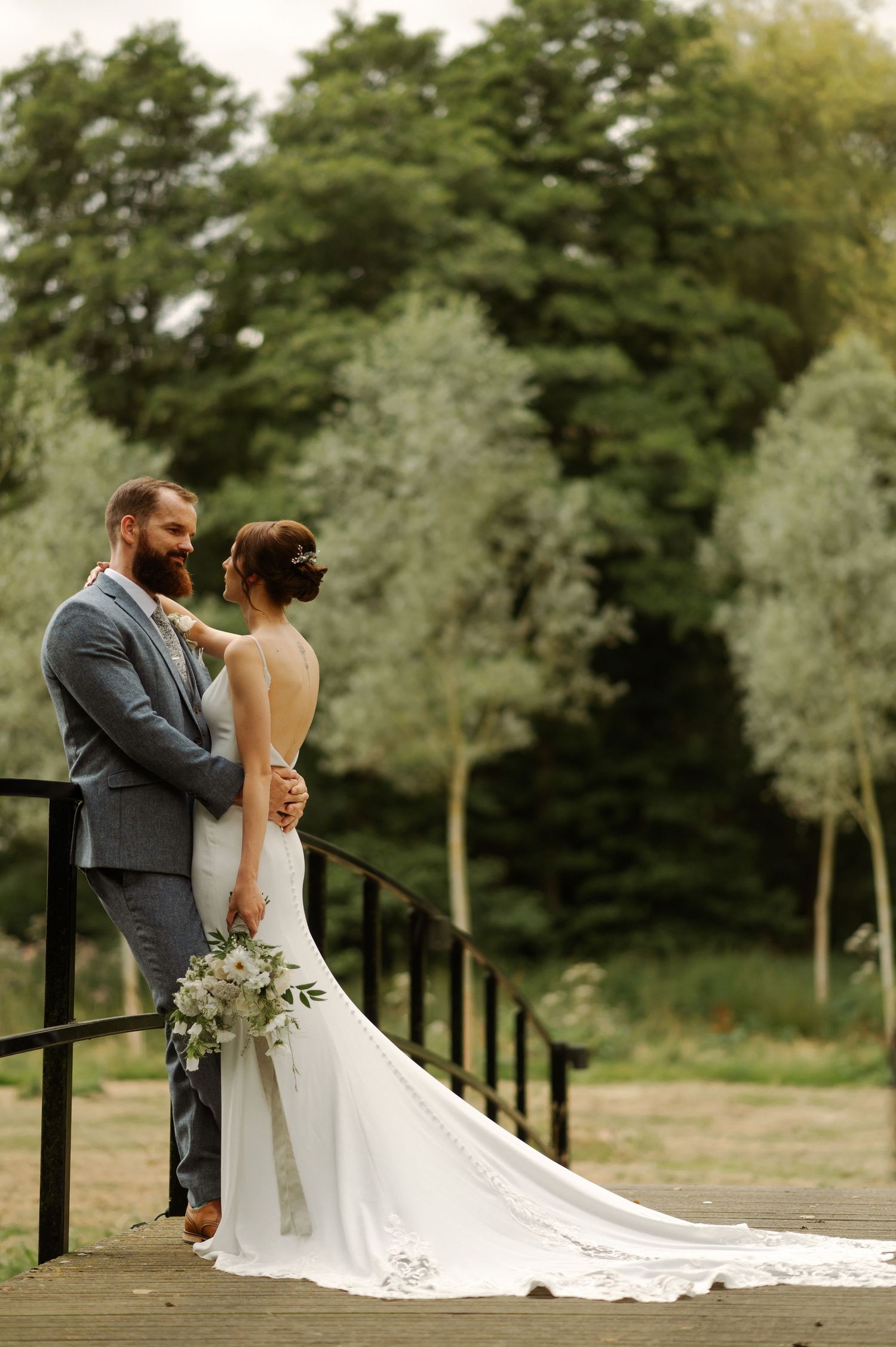 A bride and groom stand together on a wooden deck surrounded by lush green trees while her long white train flows behind.
