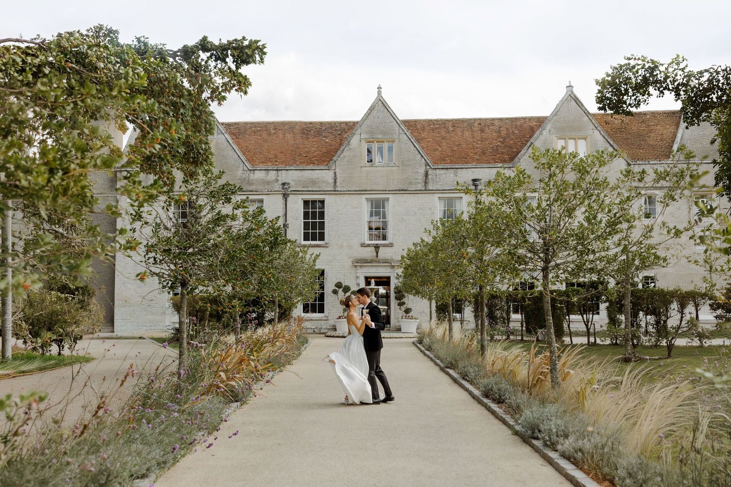 Couple sharing a romantic kiss on a tree-lined pathway in front of an elegant white wedding venue.