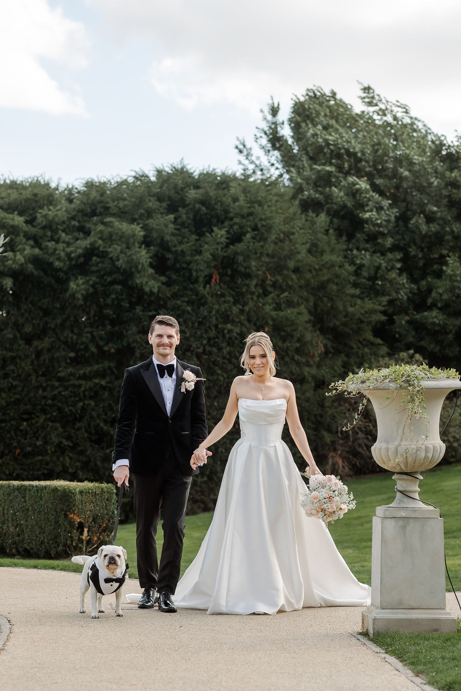 Bride and groom walk hand-in-hand with their bulldog on a manicured garden path, bride holding a white bouquet.
