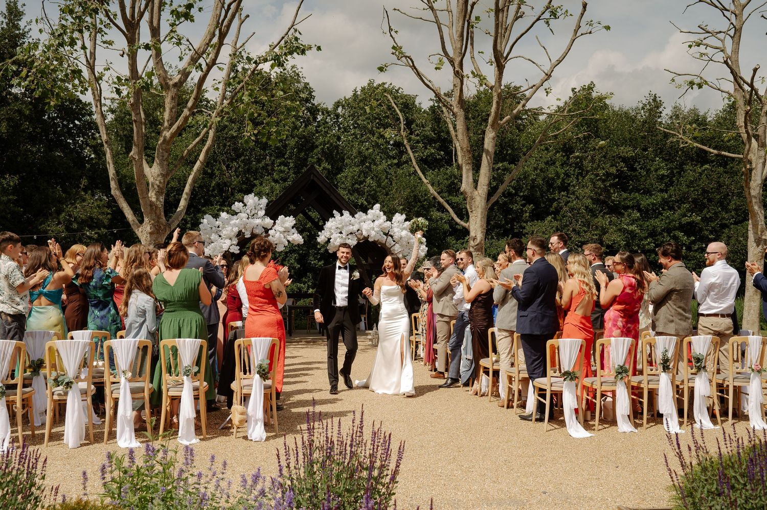 Wedding guests gather outdoors in a garden ceremony setting with trees and greenery in the background.