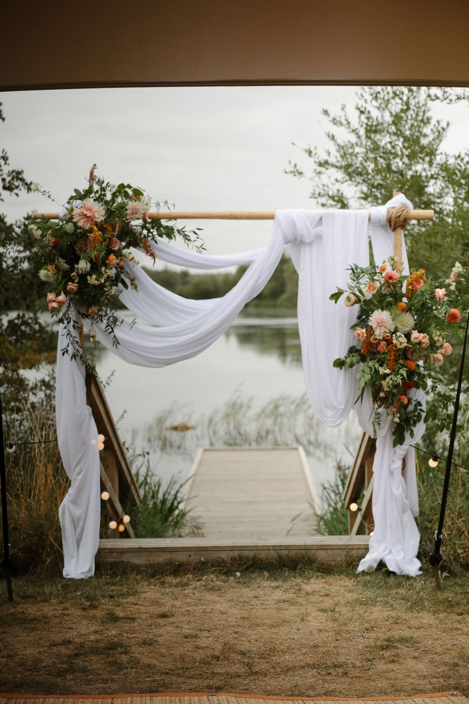 Rustic wooden wedding arch with white fabric draping and peach and orange floral arrangements overlooking a serene lake.