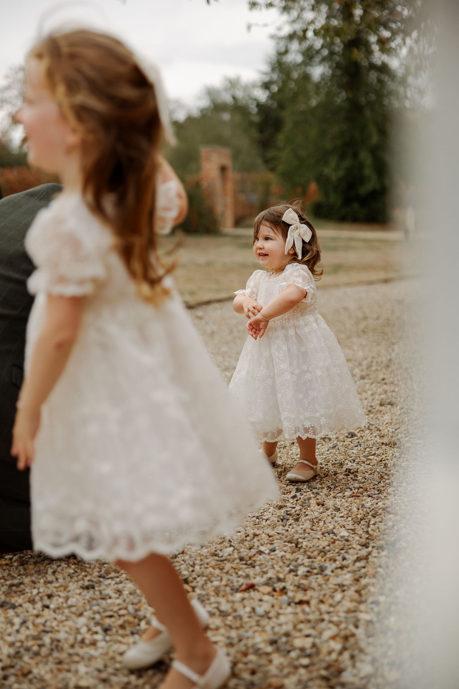 Two young flower girls in white lace dresses playing on a gravel path at a wedding venue.