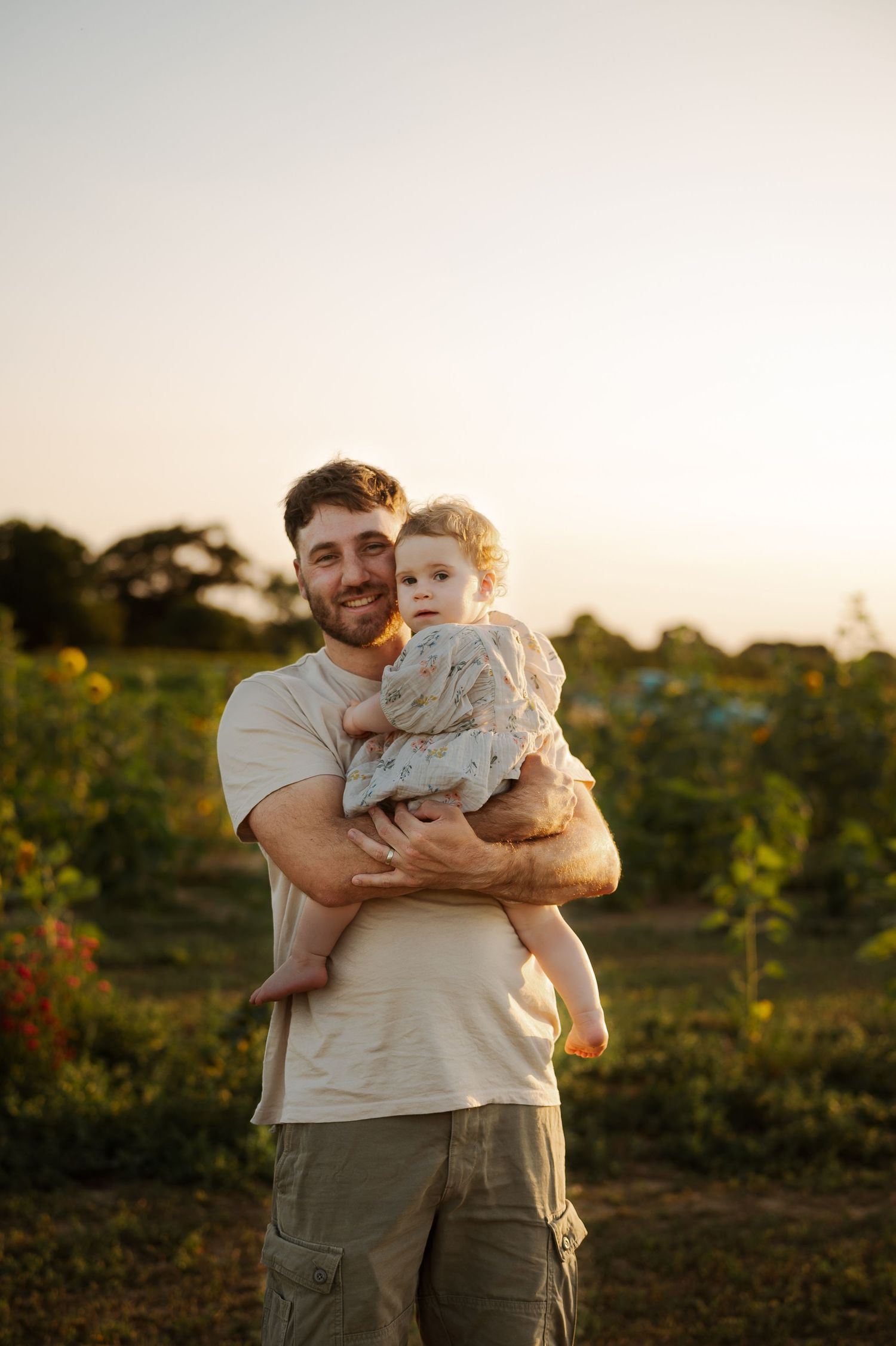 A smiling father holds his baby girl in a sunlit garden at golden hour, surrounded by lush greenery.