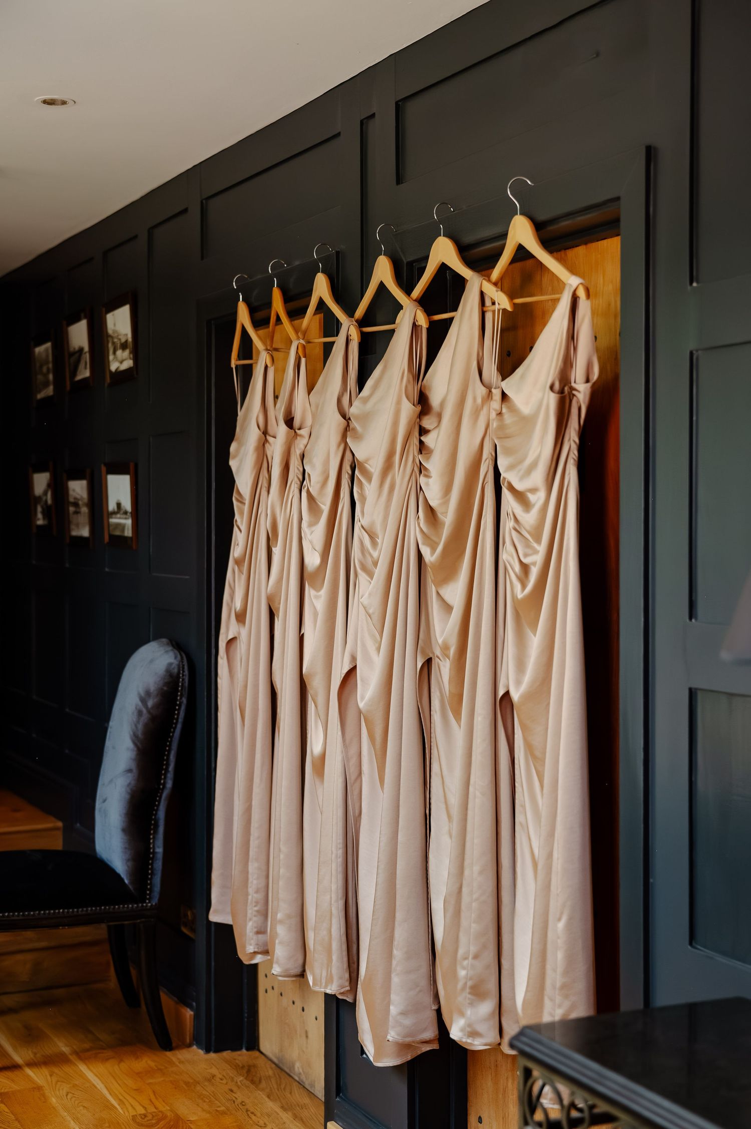 Row of blush pink bridesmaid dresses hanging on wooden hangers against a dark green wall in a bridal suite.
