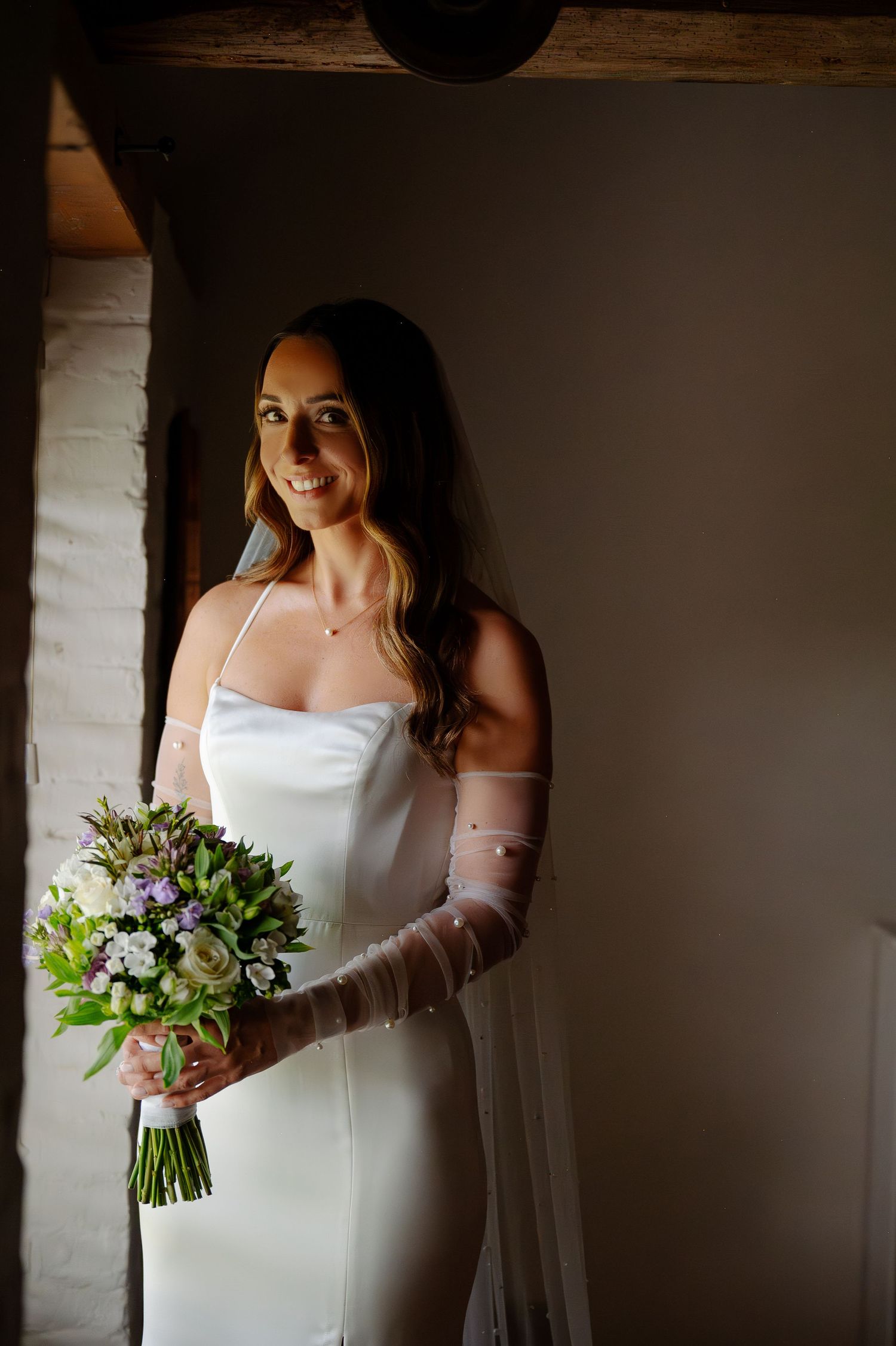 Elegant bride in sleek white gown with sheer long gloves holds a beautiful mixed floral bouquet in natural window light.