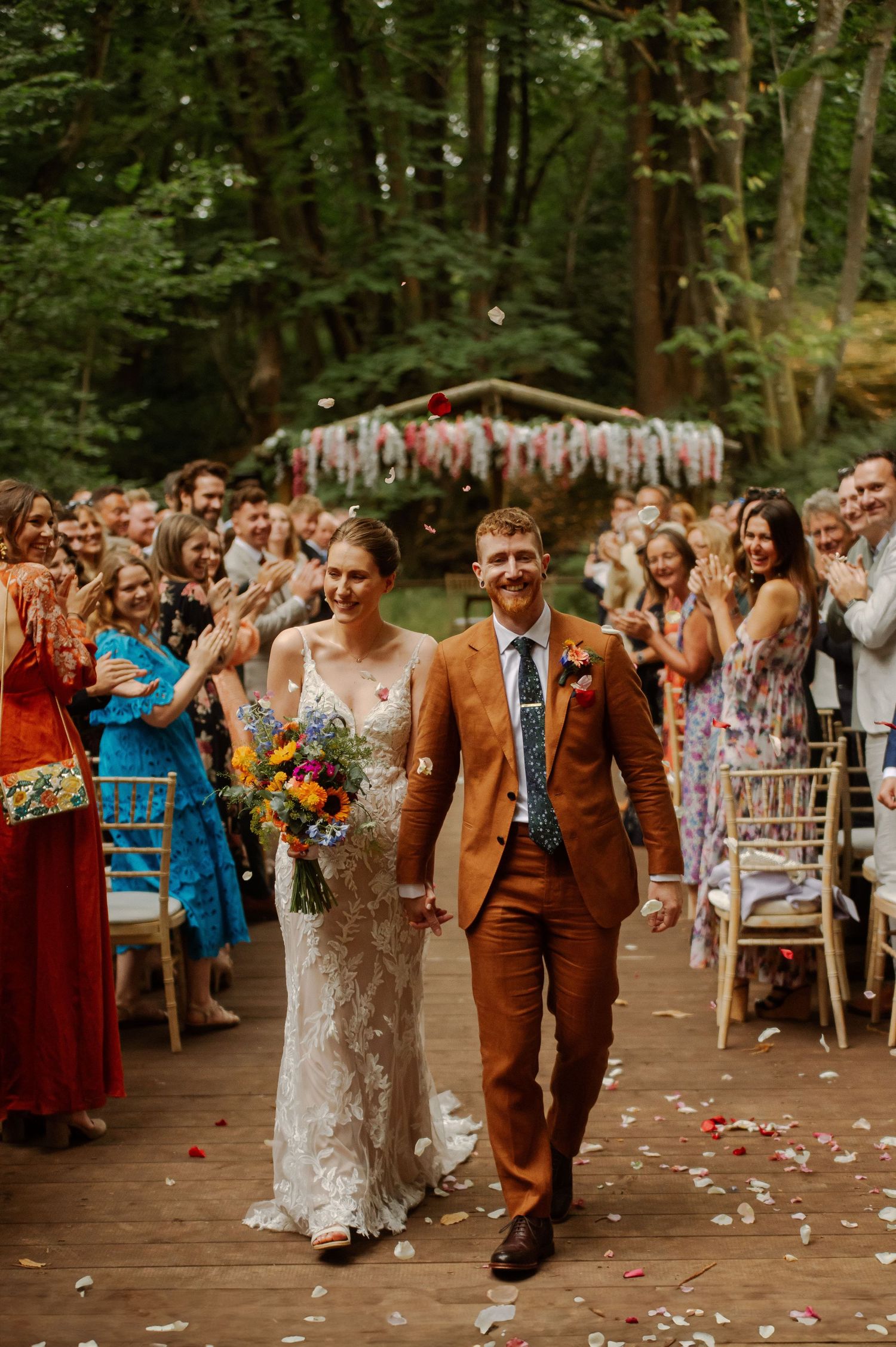 A couple walks down a wooded aisle adorned with streamers while wedding guests cheer during an outdoor forest ceremony.