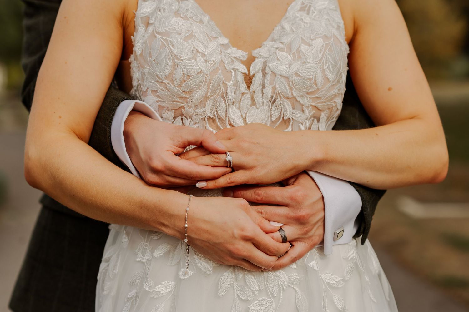 Bride and groom embrace showing wedding rings, bride wearing elegant lace gown with floral applique details.