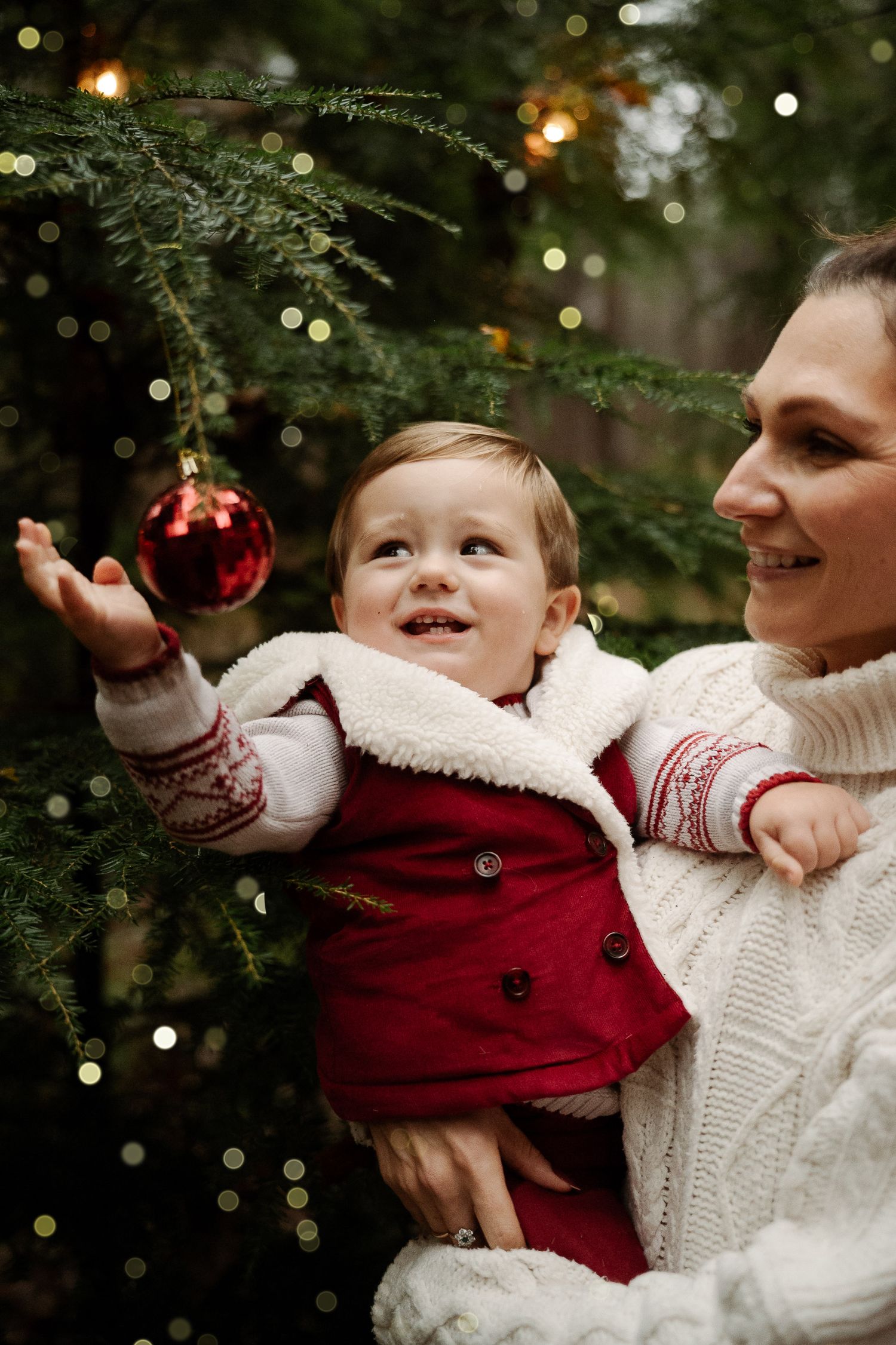 Smiling mother in white sweater holds baby in red outfit reaching for red Christmas ornament on decorated tree.