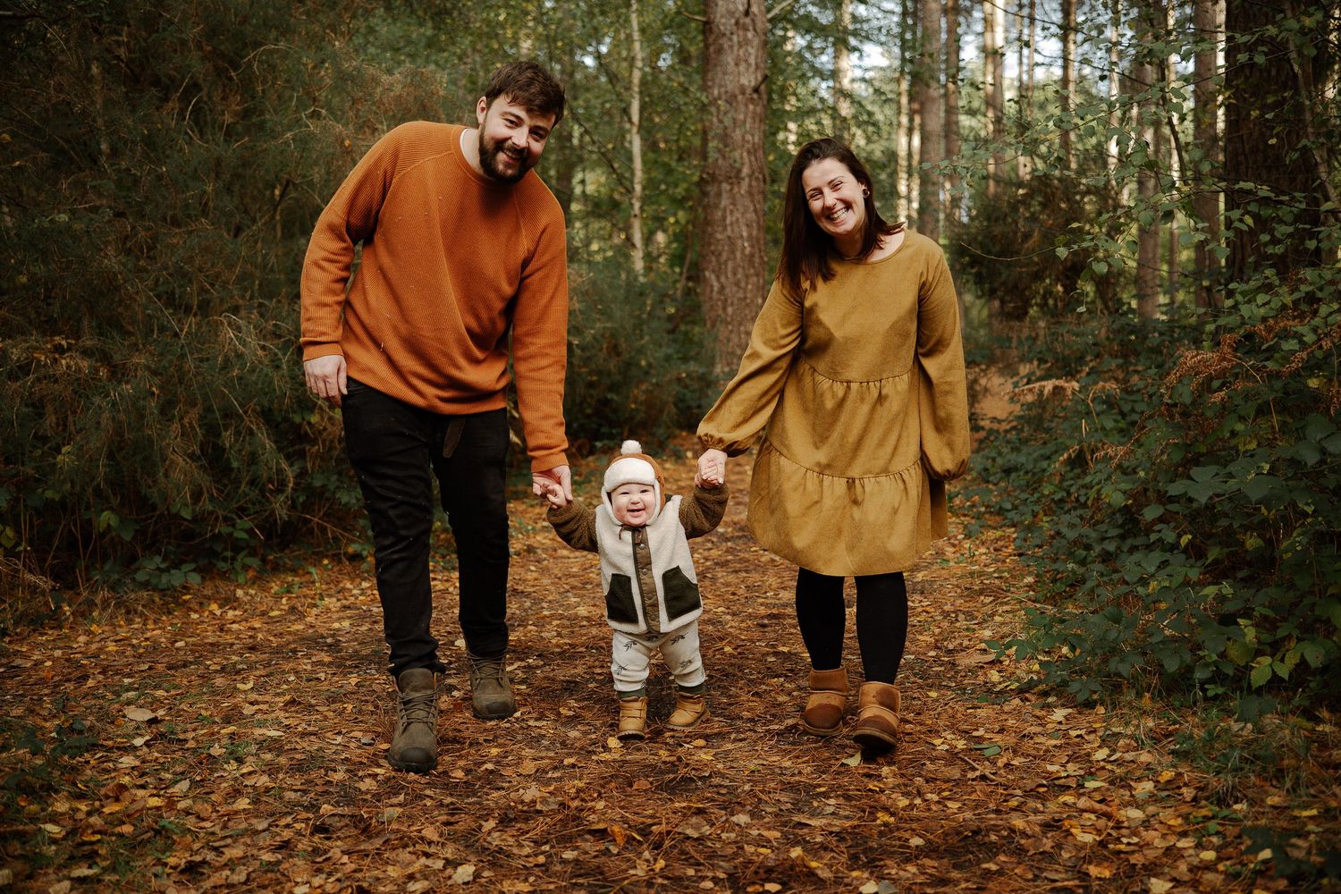 A couple in autumn attire walks with a toddler on a leaf-covered forest path during fall season.
