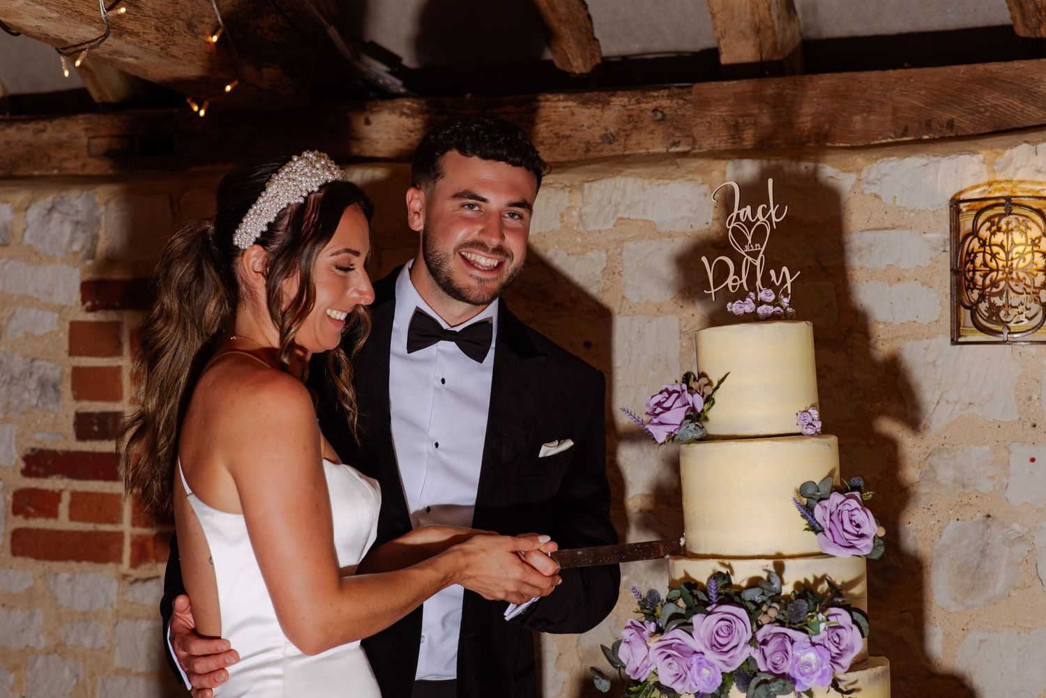 Elegant wedding reception setup with purple floral centerpieces and rustic stone wall backdrop.