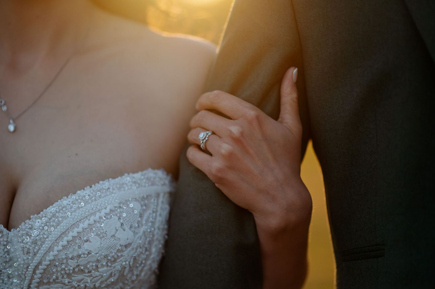 Bride in beaded gown showing diamond engagement ring while holding groom's arm in golden sunset light.