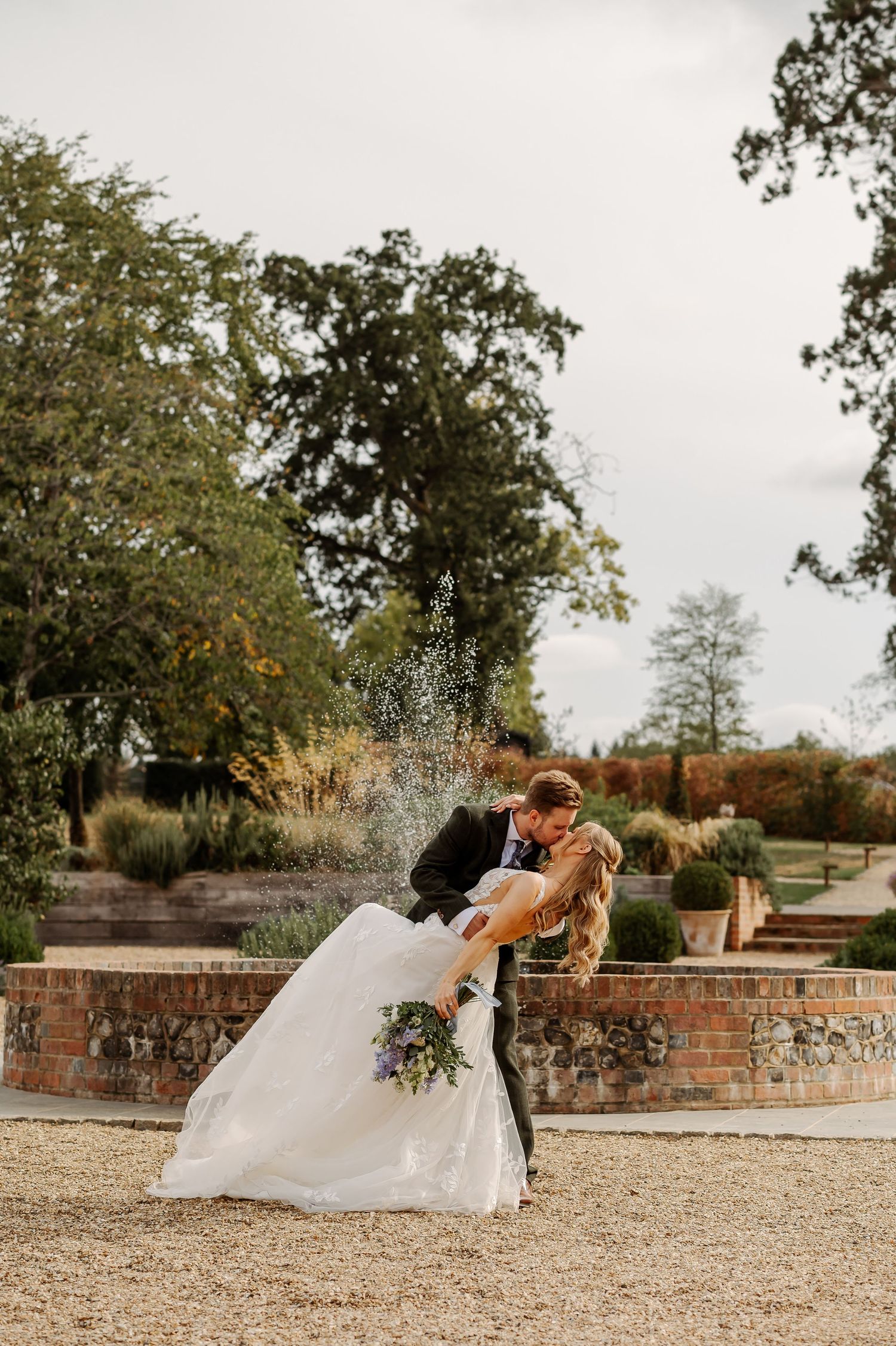 Bride and groom share a romantic dip kiss in front of a garden fountain, bride holding a purple bouquet.