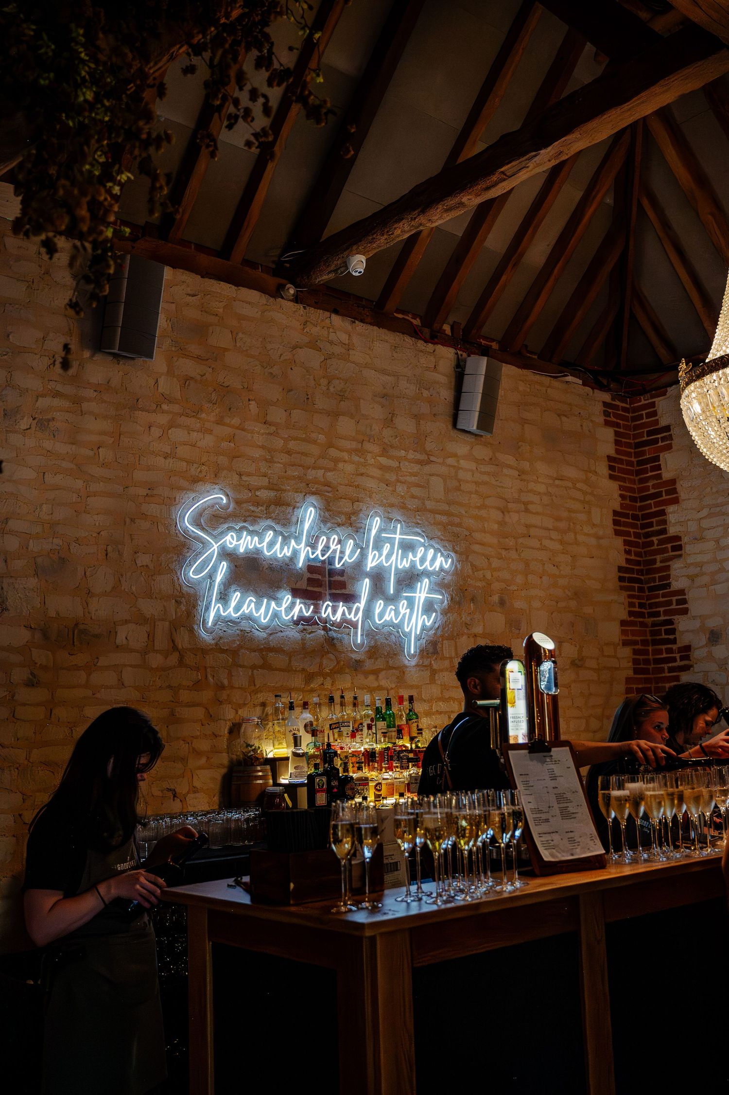 Neon sign glows above a rustic brick bar with bottles and glassware displayed on wooden shelves.