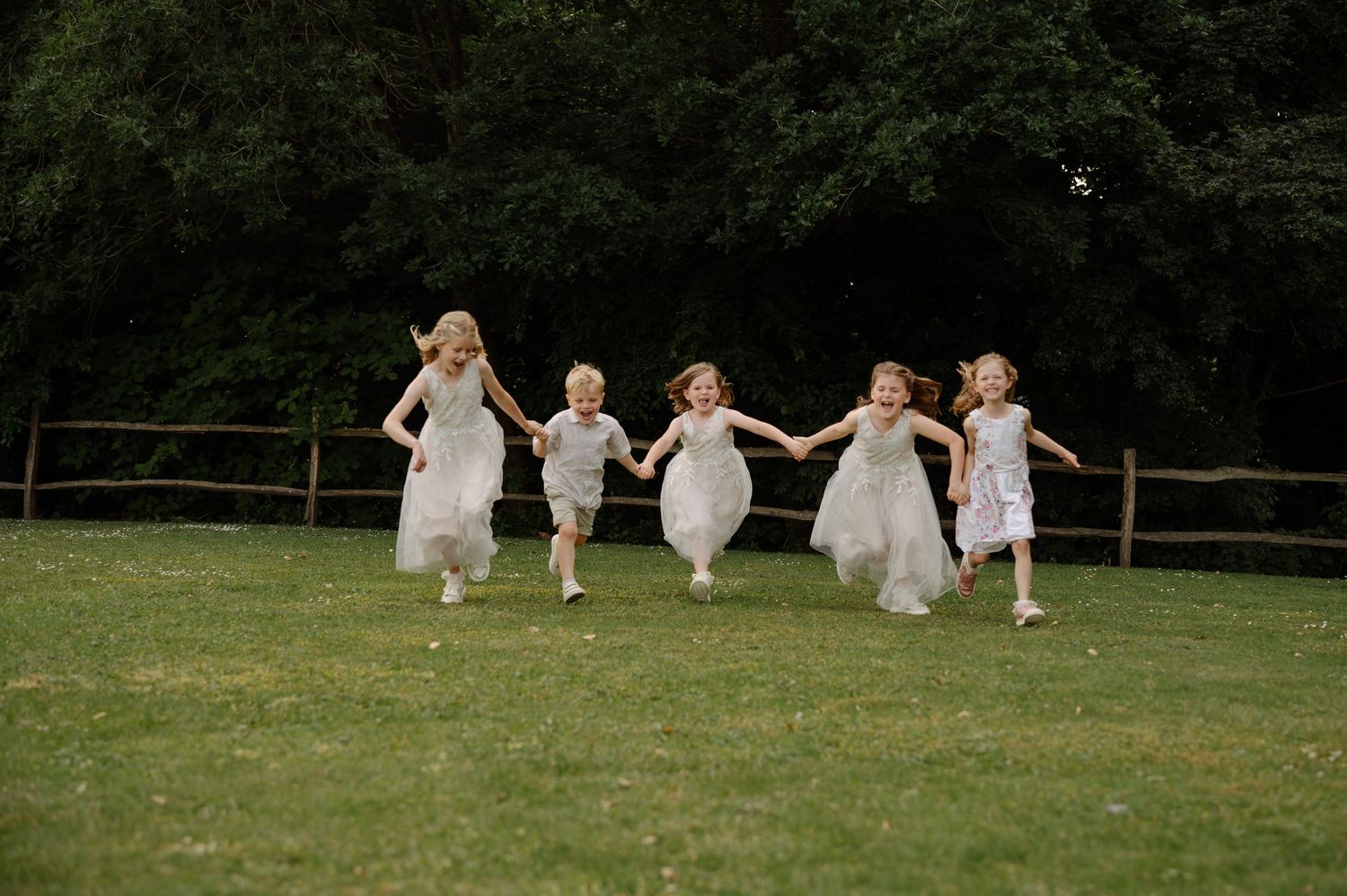Children in white dresses run and play together on a grassy lawn during an outdoor gathering.