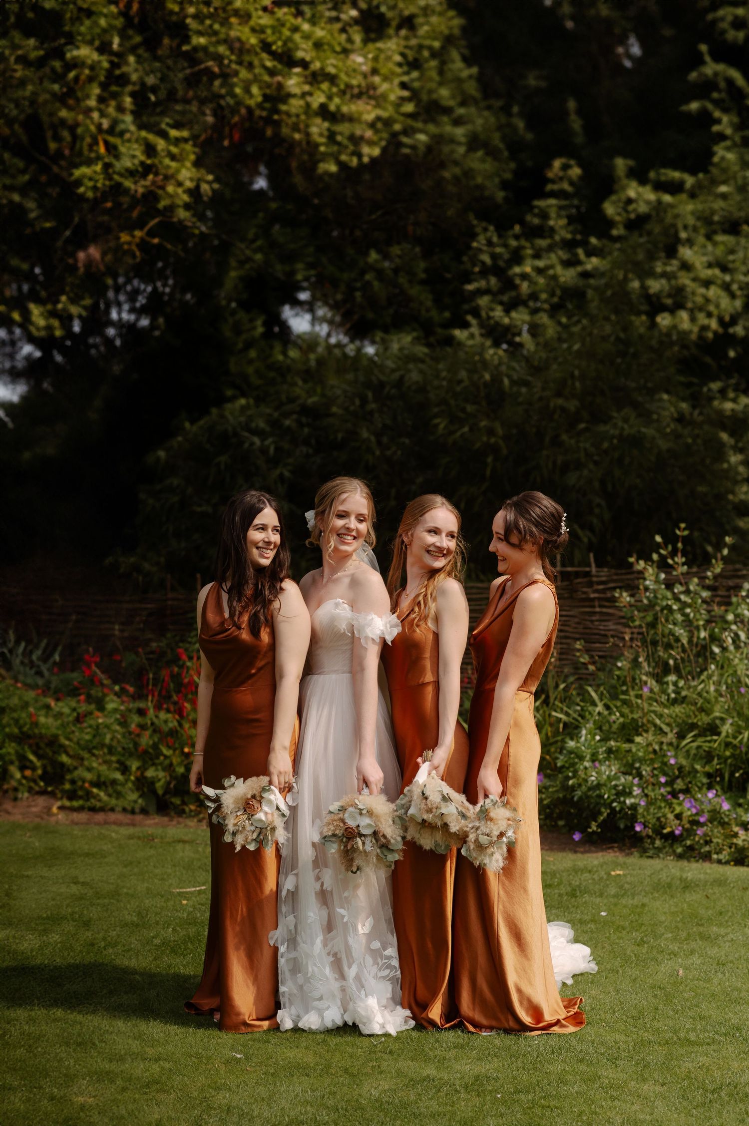 A group in coordinating autumn-toned dresses stands together on a green lawn during a wedding celebration.