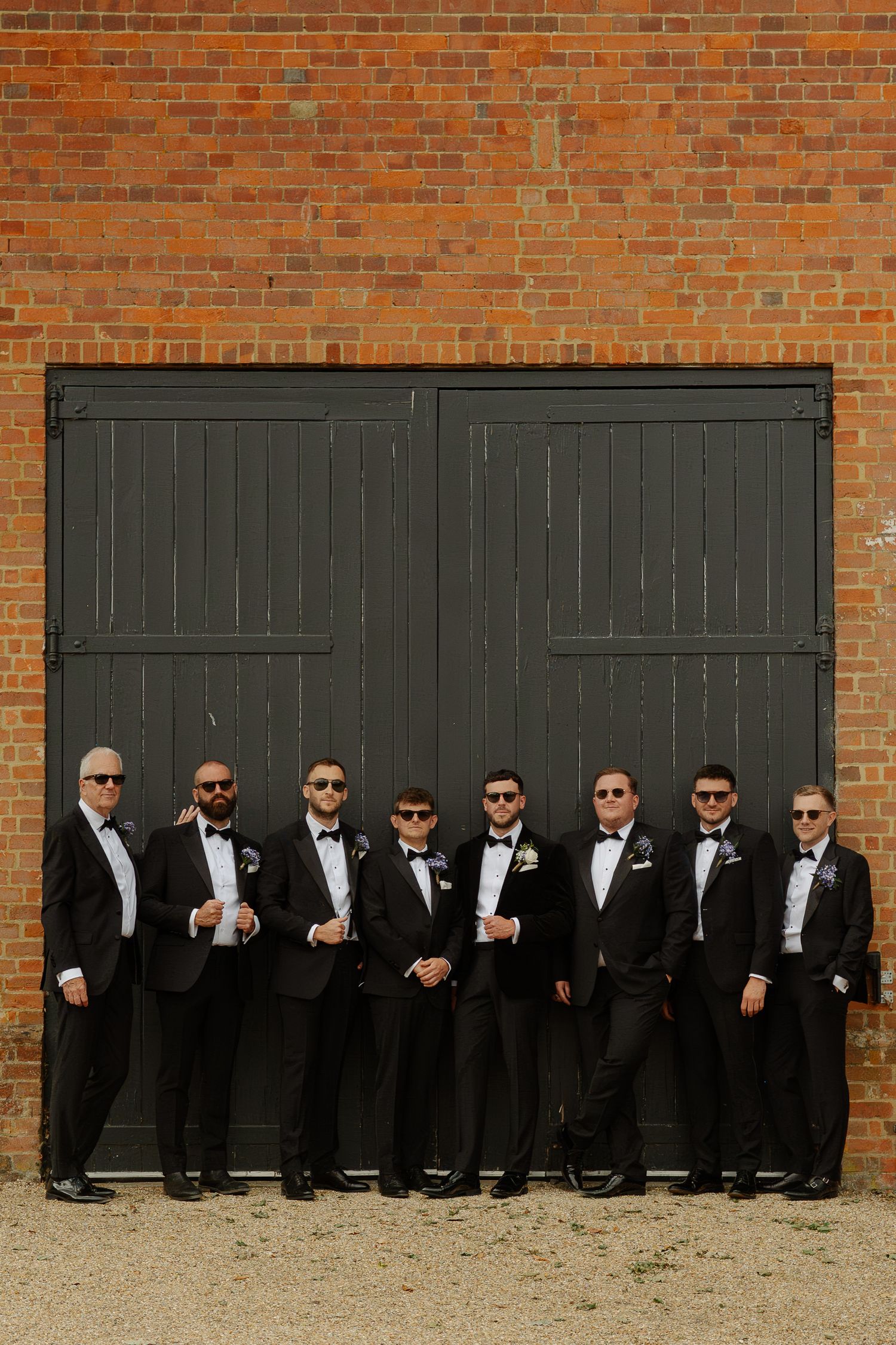 Groomsmen in black tuxedos and sunglasses pose casually against a rustic brick wall with dark wooden doors.