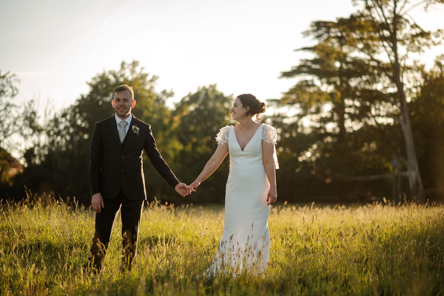 Bride and groom holding hands in golden sunlit meadow, bride in white gown, groom in dark suit, trees in background.