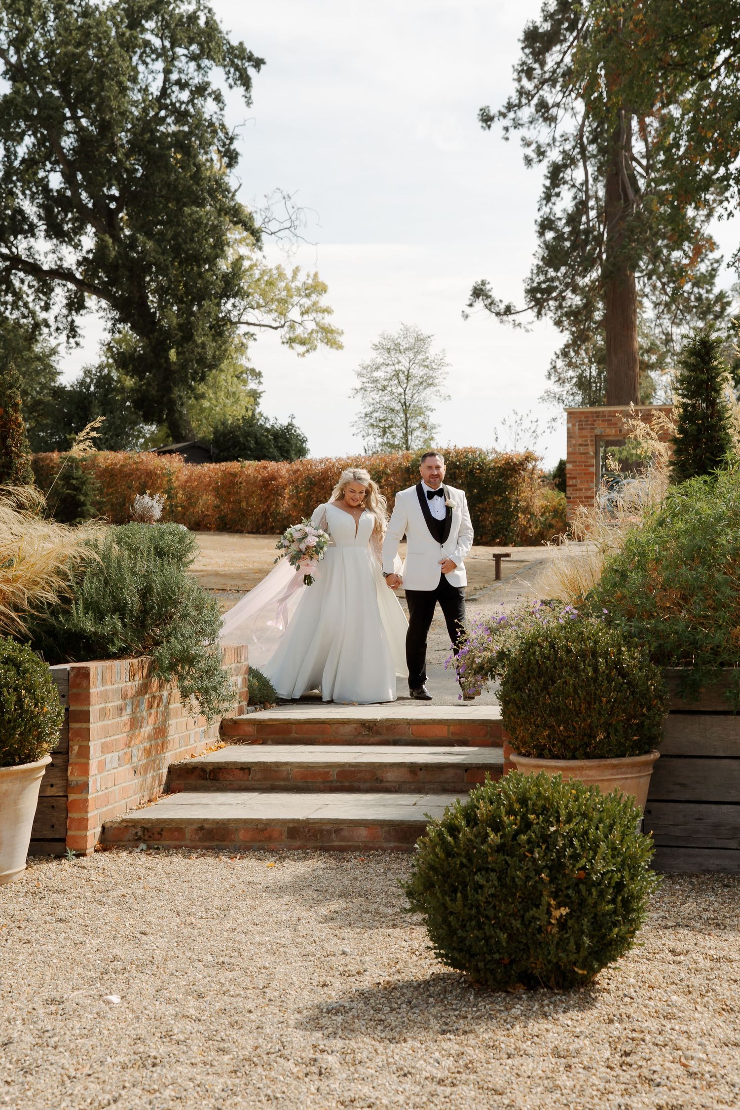 Bride and groom walk hand in hand down garden steps at an elegant outdoor wedding venue surrounded by lush greenery.