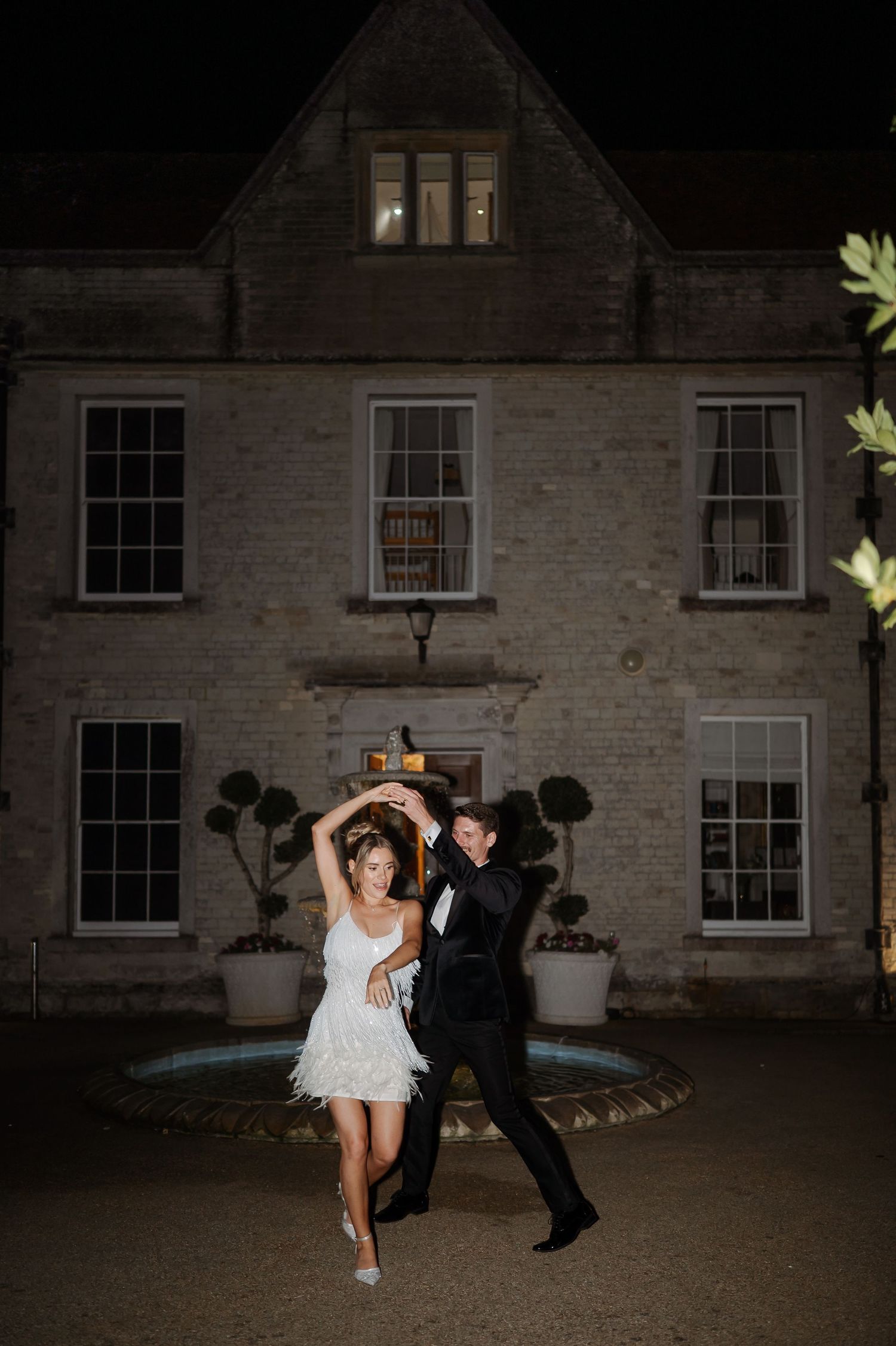 Couple dancing joyfully outside an elegant stone manor at night, woman in white fringe dress, man in black suit.