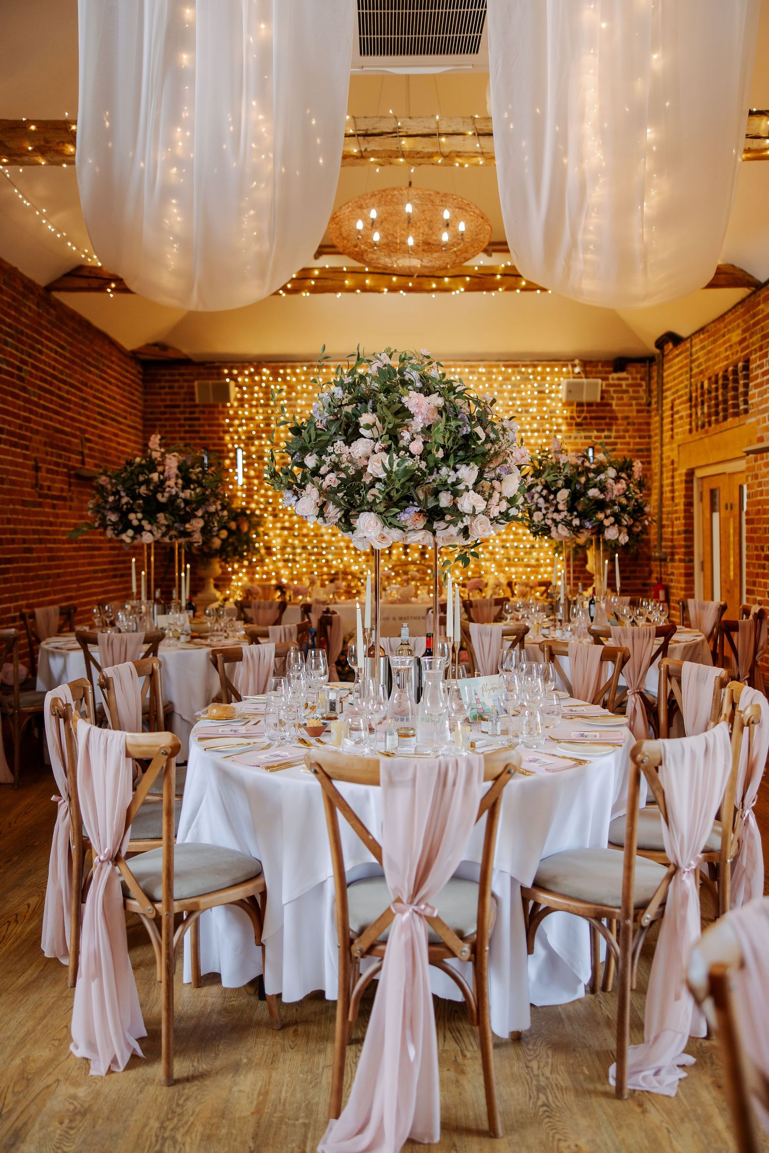 Elegant wedding reception venue with pink chair sashes, tall floral centerpieces, fairy lights, and white draping.