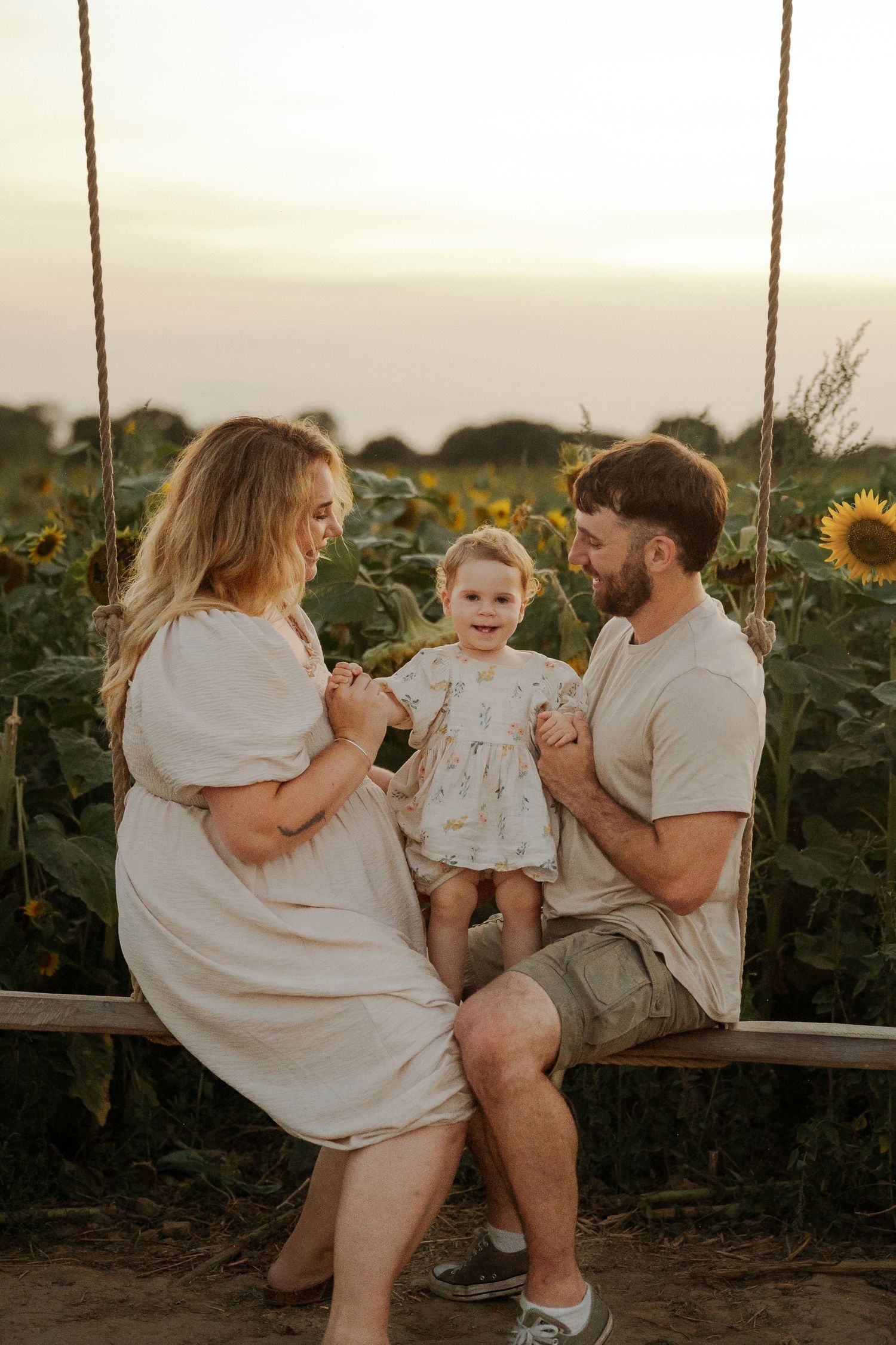 Family of three sitting on a swing in a sunflower field at golden hour, parents smiling at their baby girl.