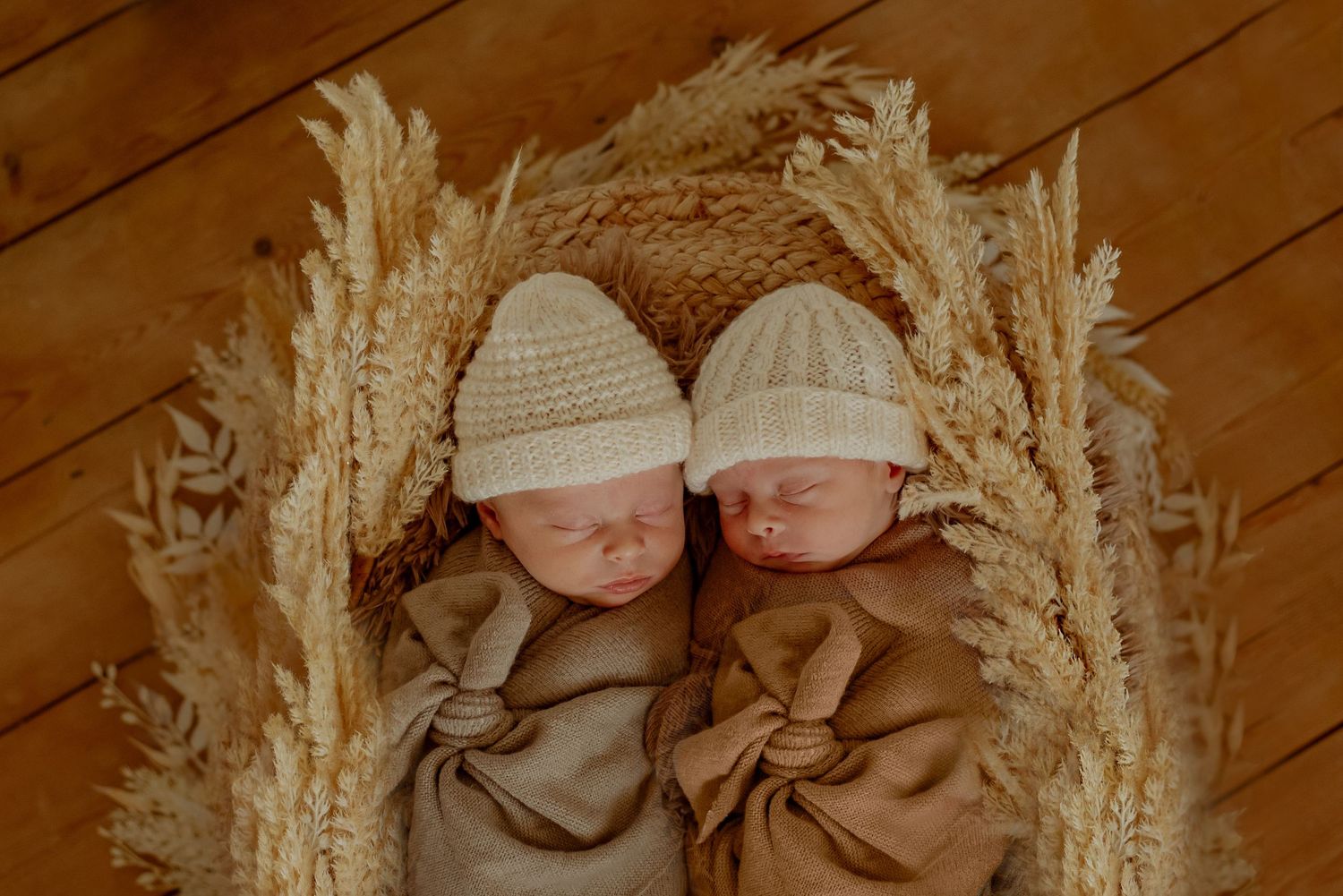 Two sleeping newborn babies wearing white knit caps lie together on wooden floor surrounded by dried pampas grass.