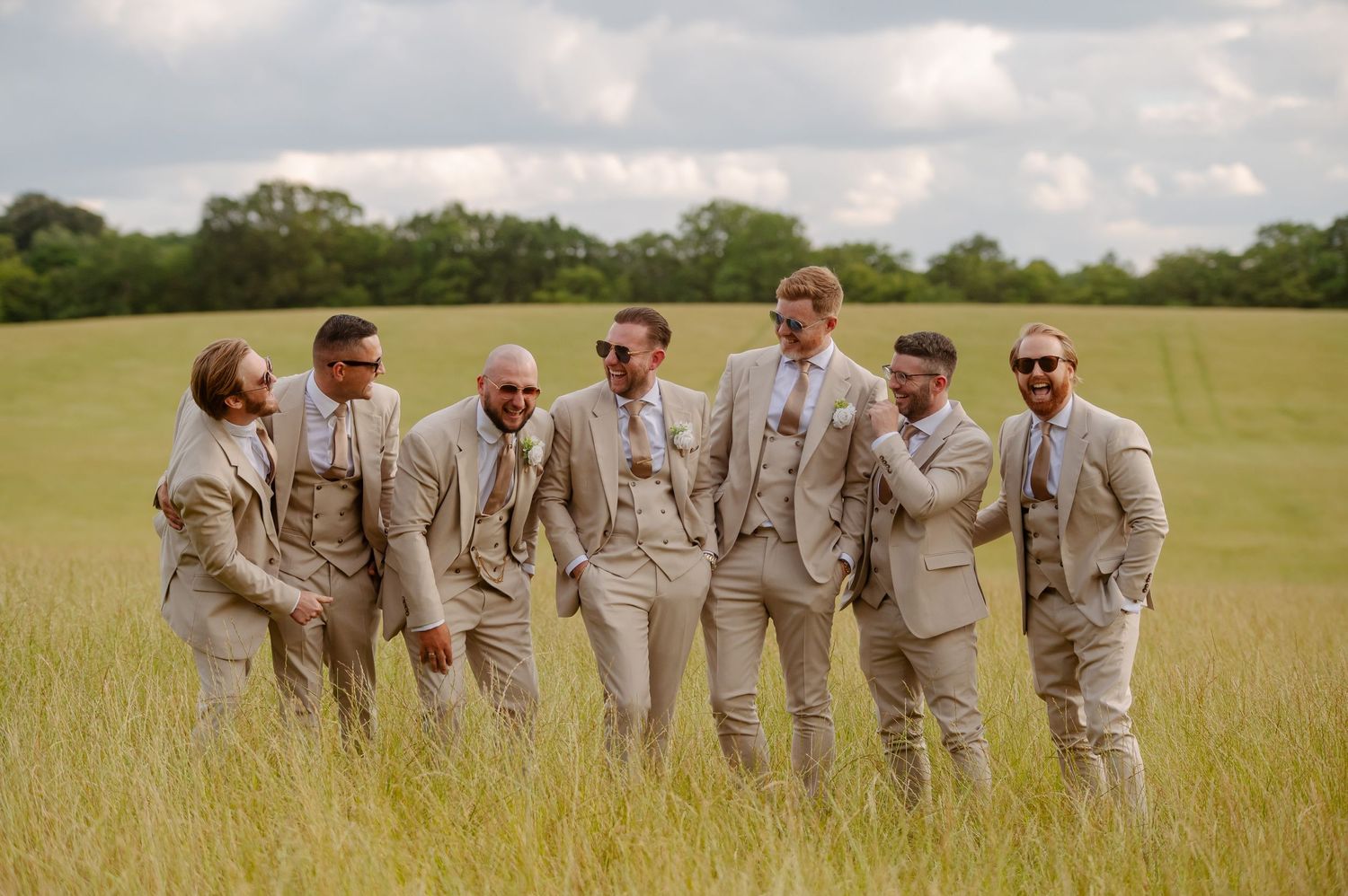 Groomsmen in light beige suits laugh together while standing in sunny meadow.