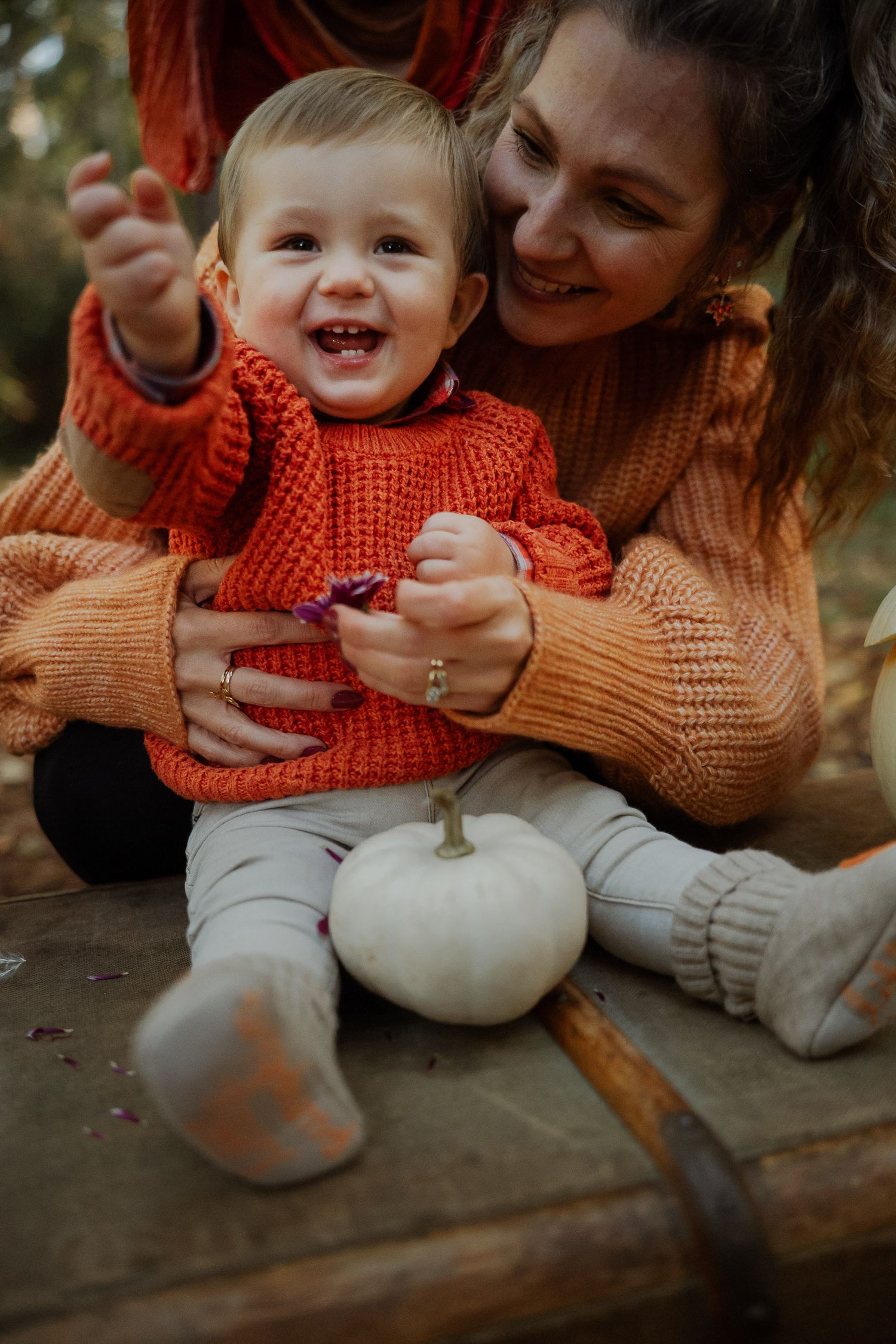 A joyful toddler in an orange knit sweater sits with a white pumpkin while a smiling woman hugs him from behind.