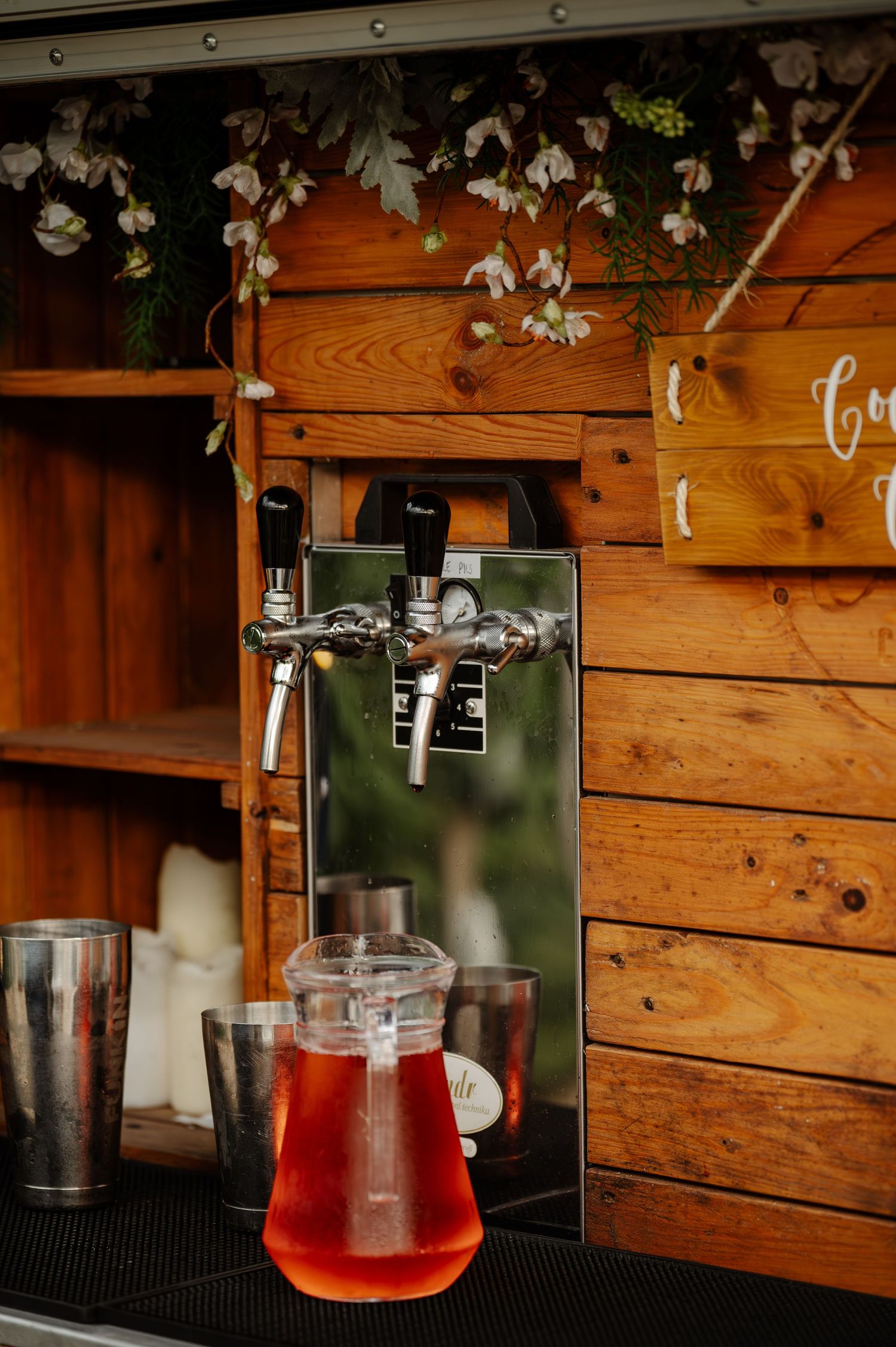 A wooden bar display with hanging decorations and red cocktails in glass containers.
