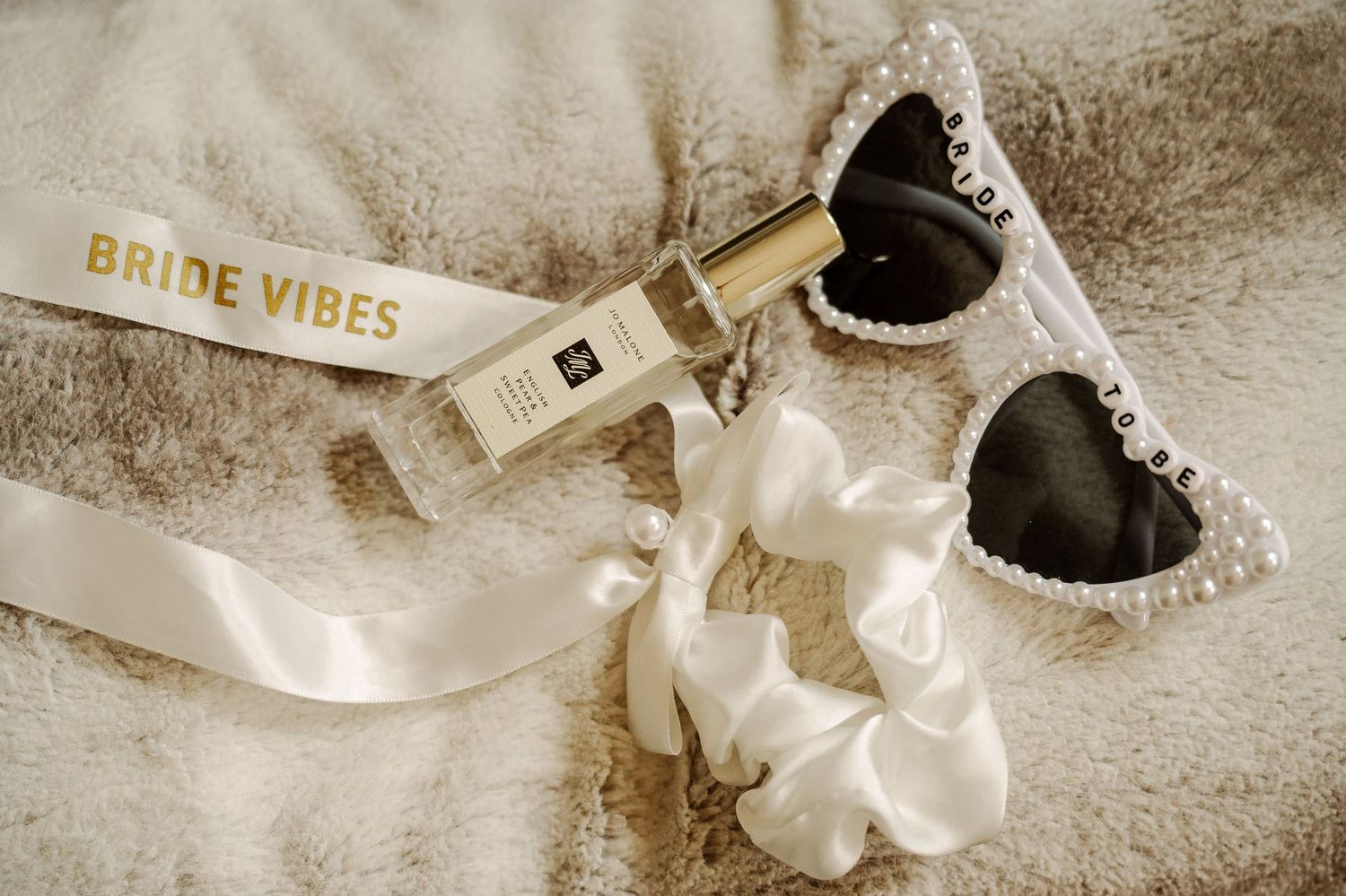 Wedding accessories including white shoes, pearls, and champagne bottle arranged on textured beige background.