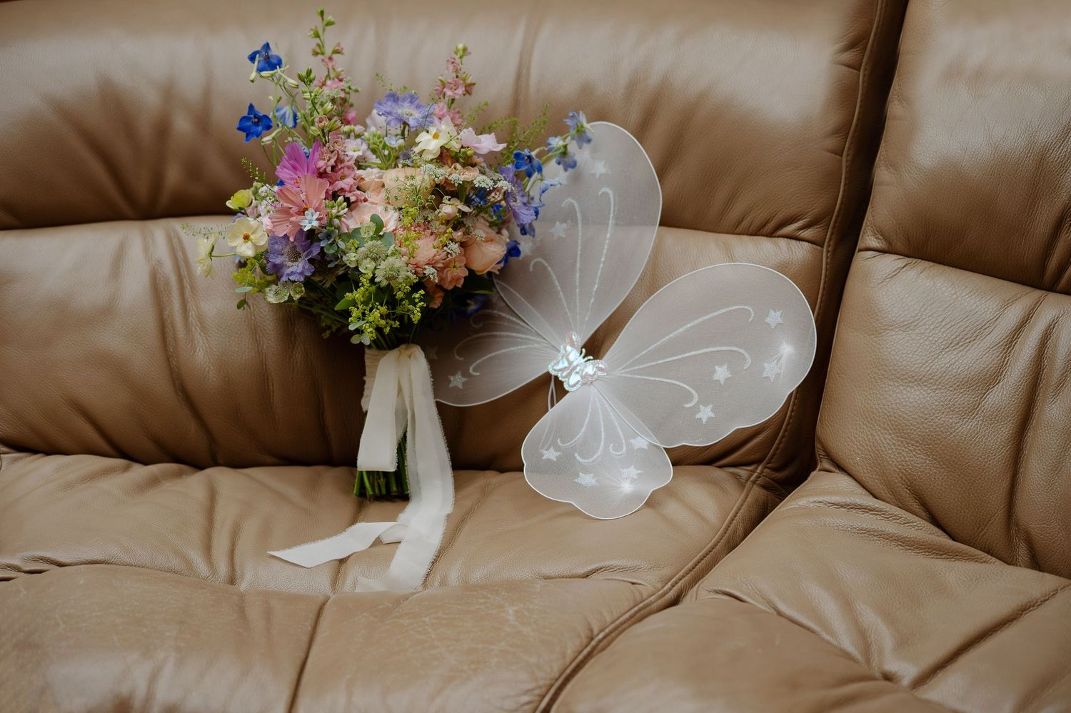 A delicate floral bouquet with pink and blue wildflowers sits next to white fairy wings on a tan leather couch.