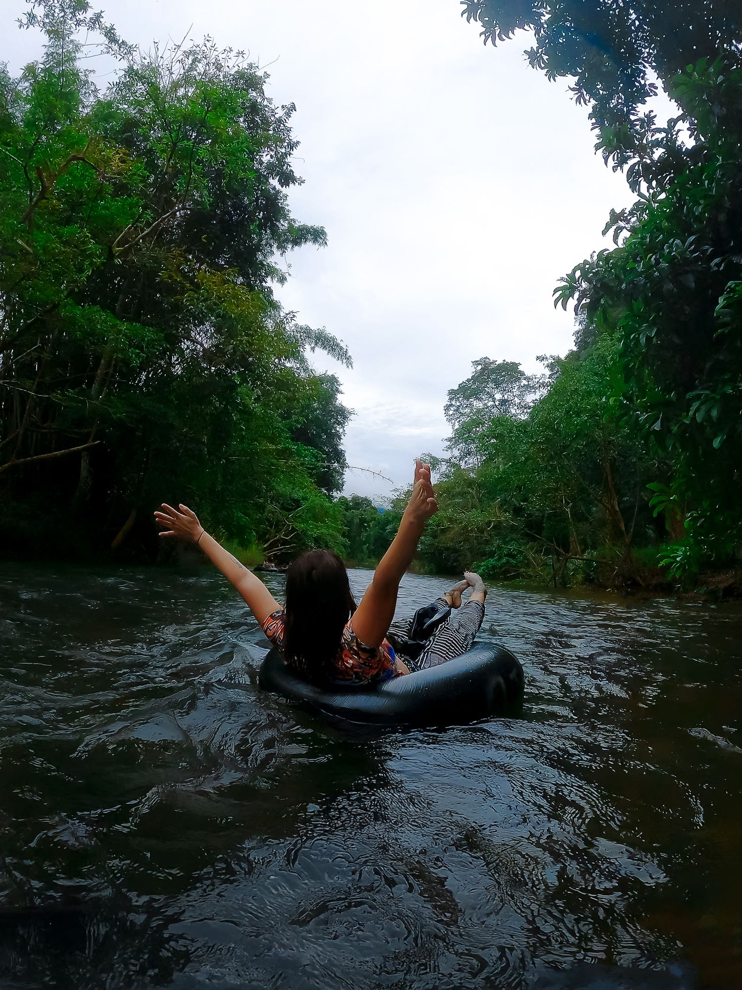 Someone tubing down a river surrounded by lush green trees raises their arms in celebration while floating downstream.