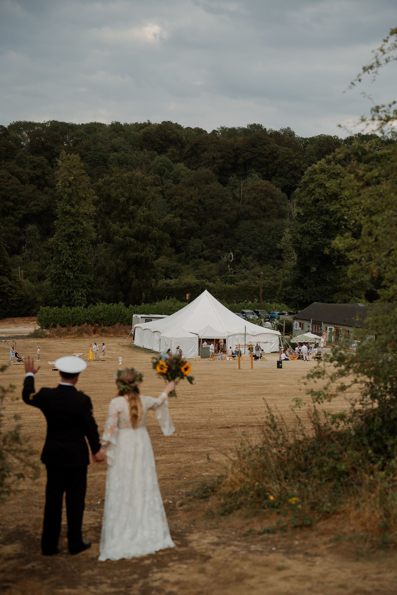 Bride and groom holding hands overlooking outdoor wedding reception tent surrounded by lush green trees on a cloudy day.