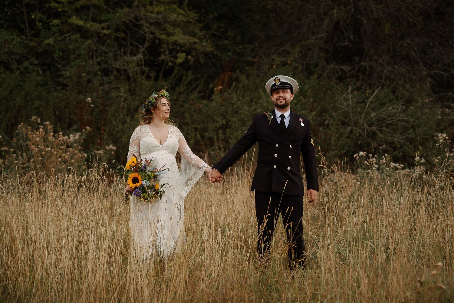 Bride and groom holding hands in golden field, she holds sunflowers, he wears navy uniform with captain hat.