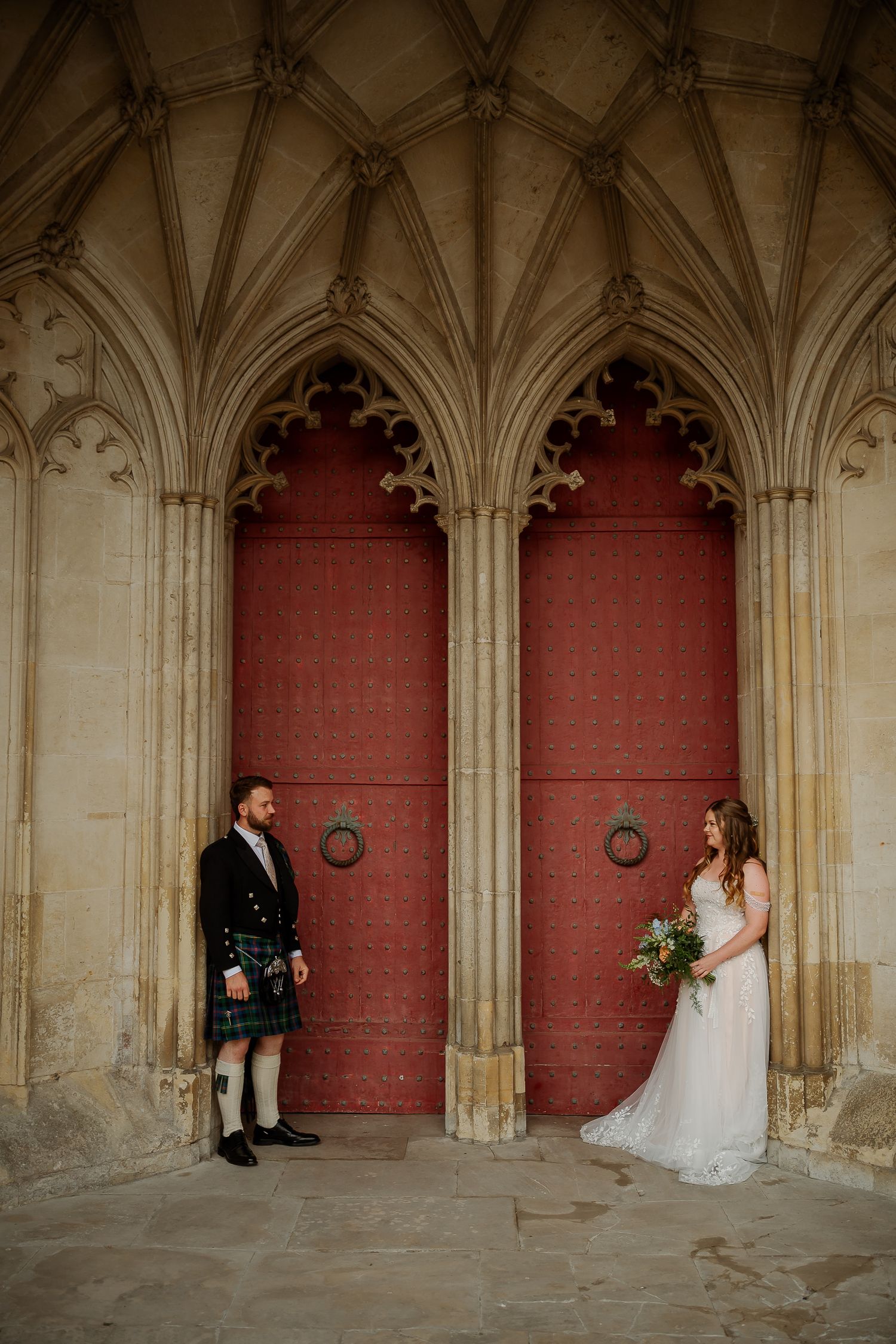 Bride and groom pose before grand Gothic red doors, he in tartan kilt, she in white gown holding bouquet.