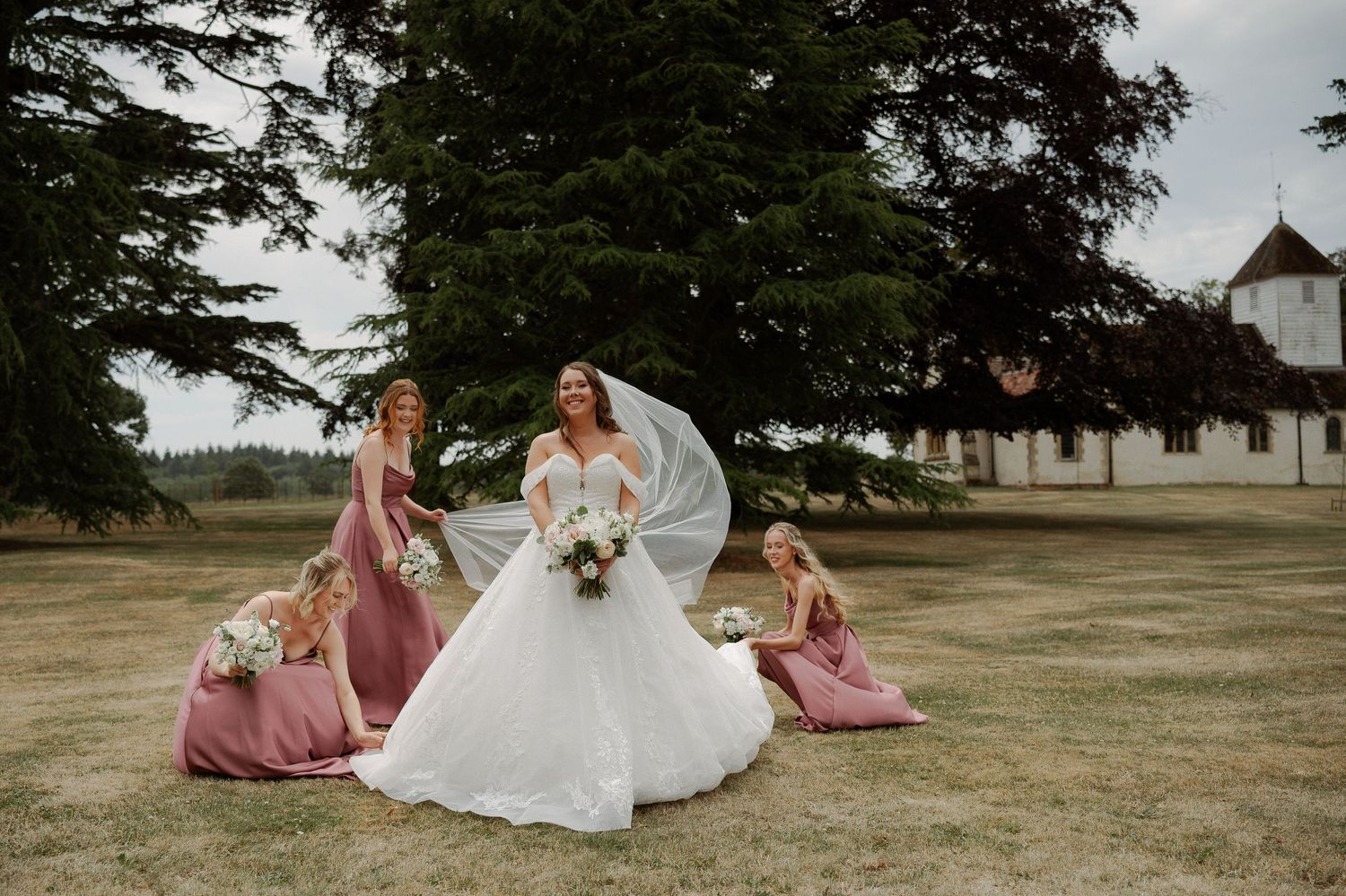 A wedding party in mauve dresses and a white gown pose together on a grassy lawn near trees.