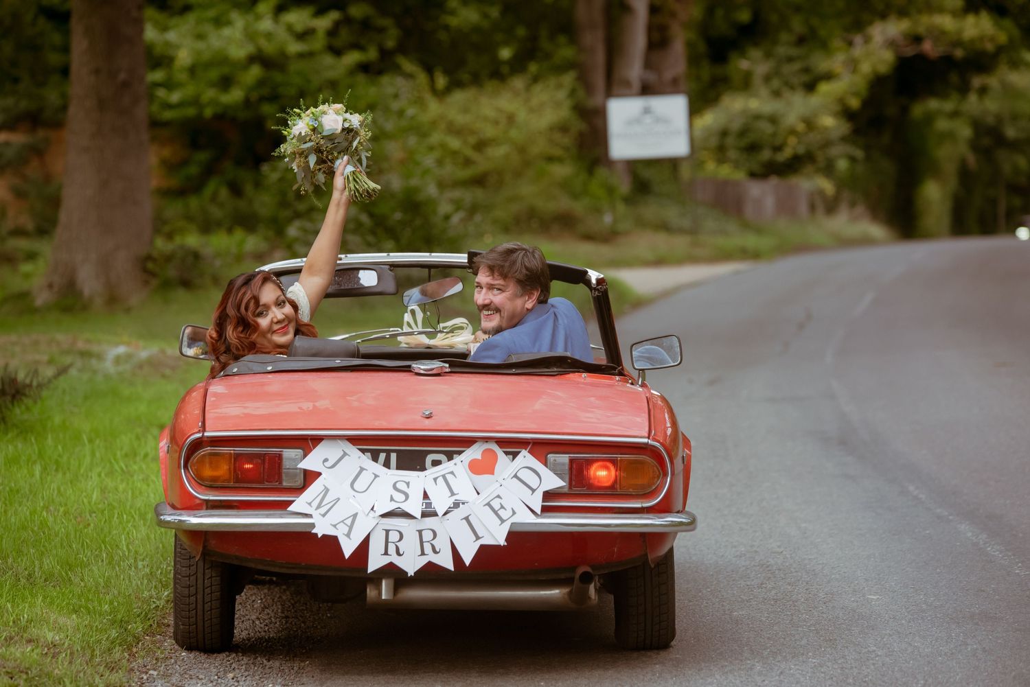 A newlywed couple driving away in a vintage red convertible decorated with wedding streamers.