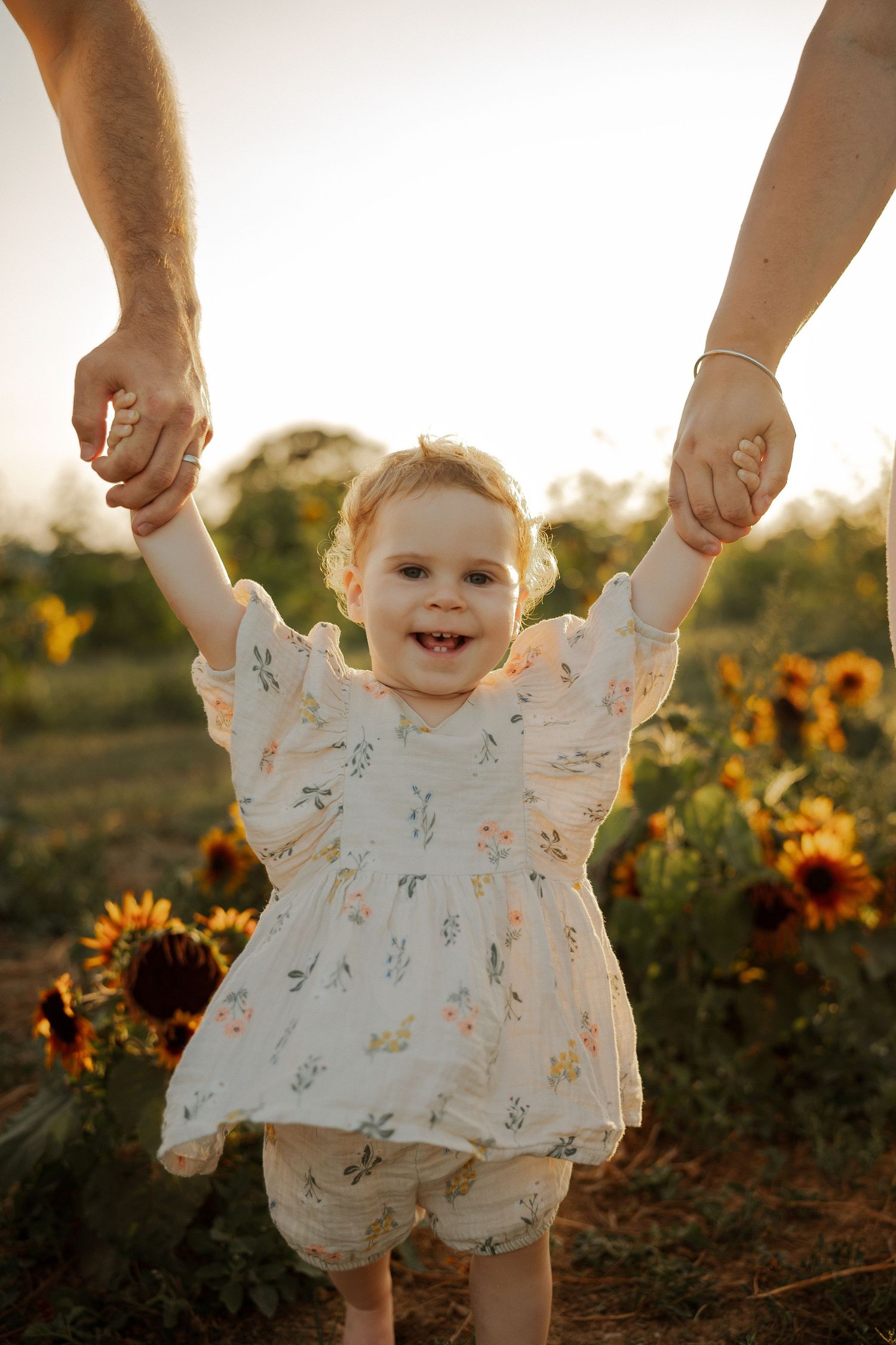 Smiling baby girl in floral dress held by parents' hands in a sunflower field at golden hour.