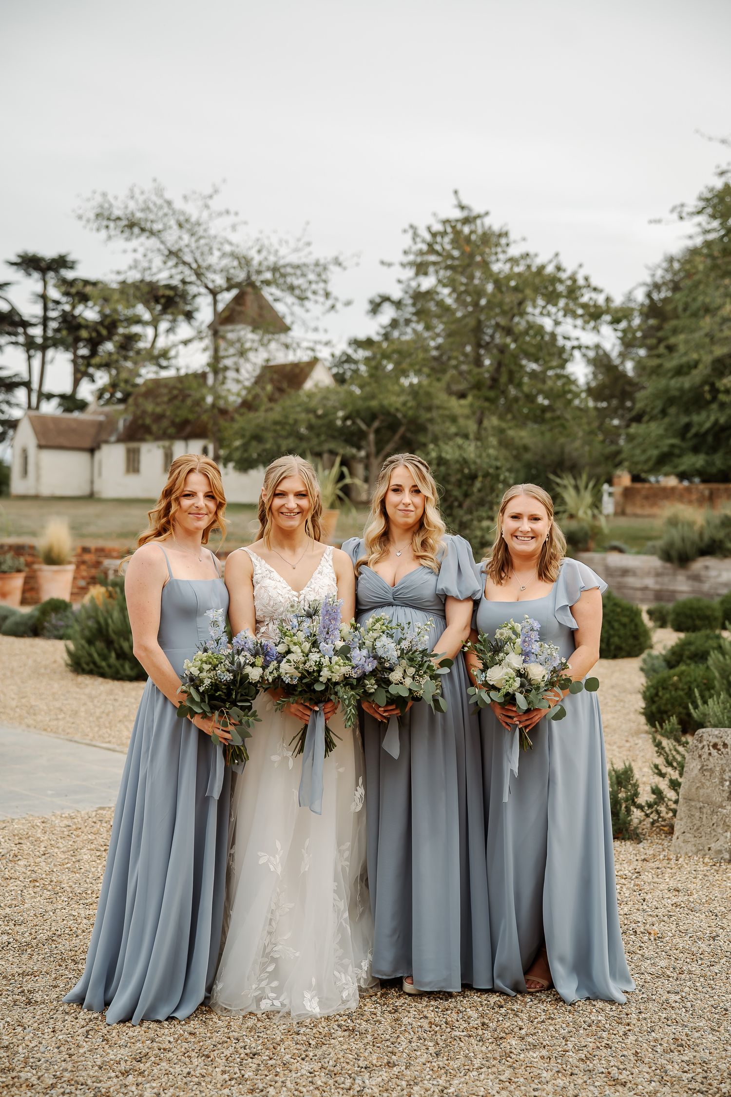 Bride in white lace gown with three bridesmaids in dusty blue dresses holding floral bouquets outdoors.