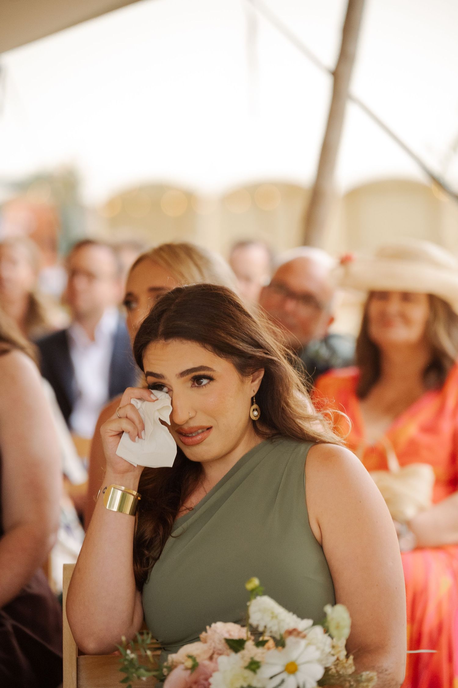 Bridesmaid in olive green dress holds white floral bouquet at wedding celebration with blurred guests in background.