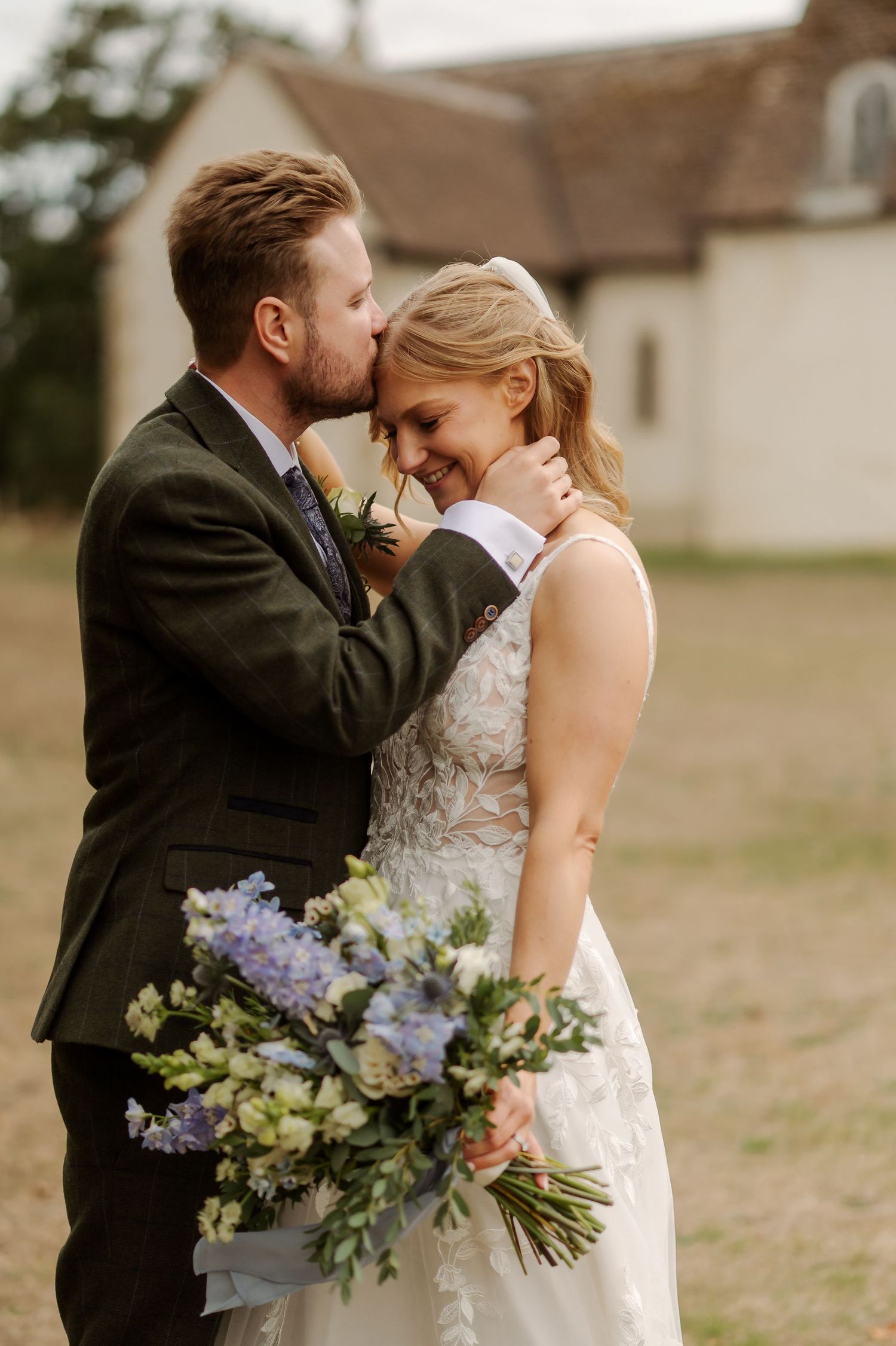 Groom in dark suit kissing bride on cheek as she smiles, holding blue and white floral bouquet outdoors.