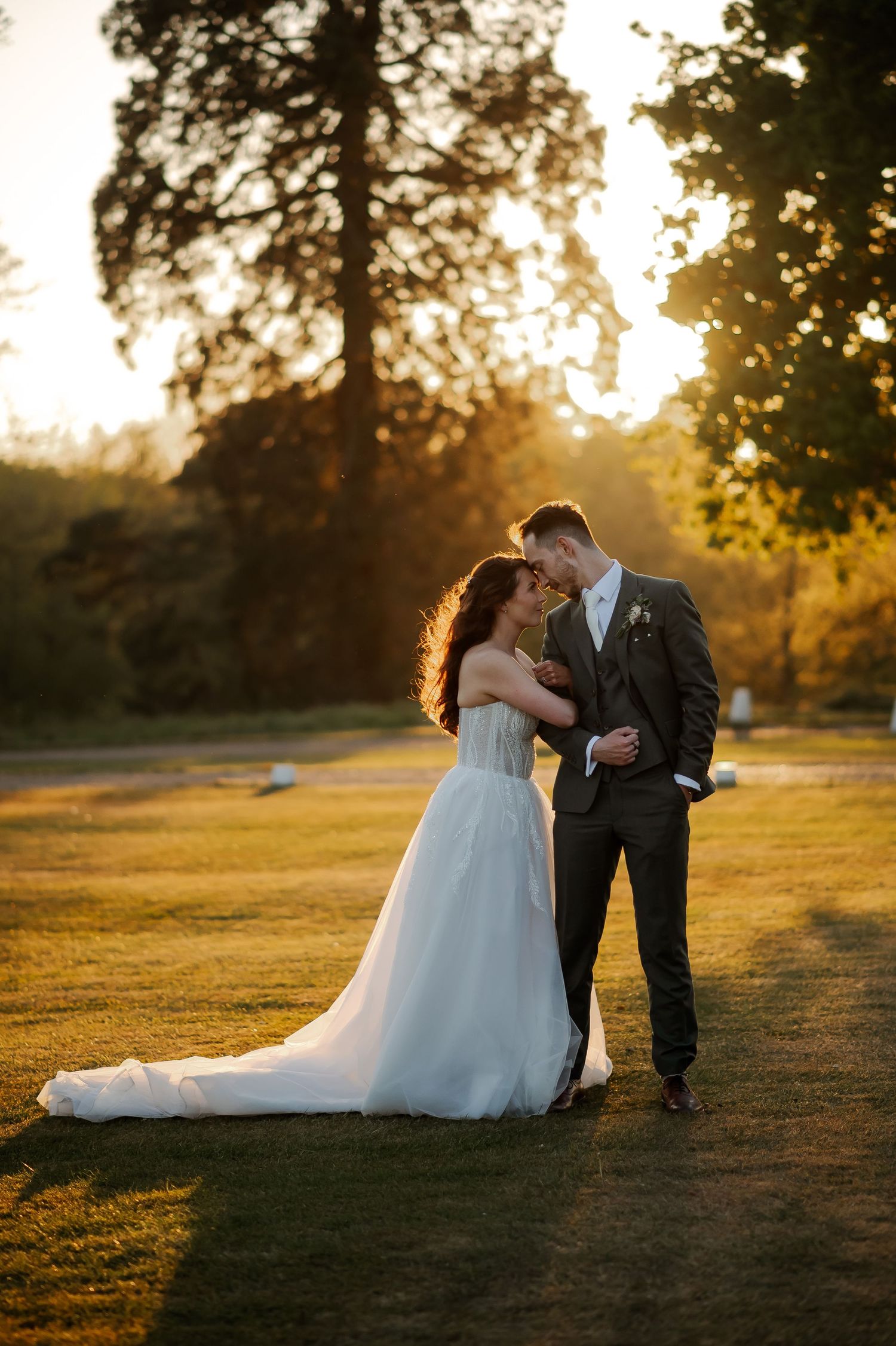 Bride and groom share a romantic sunset moment on lush green lawn, golden light filtering through tall trees.