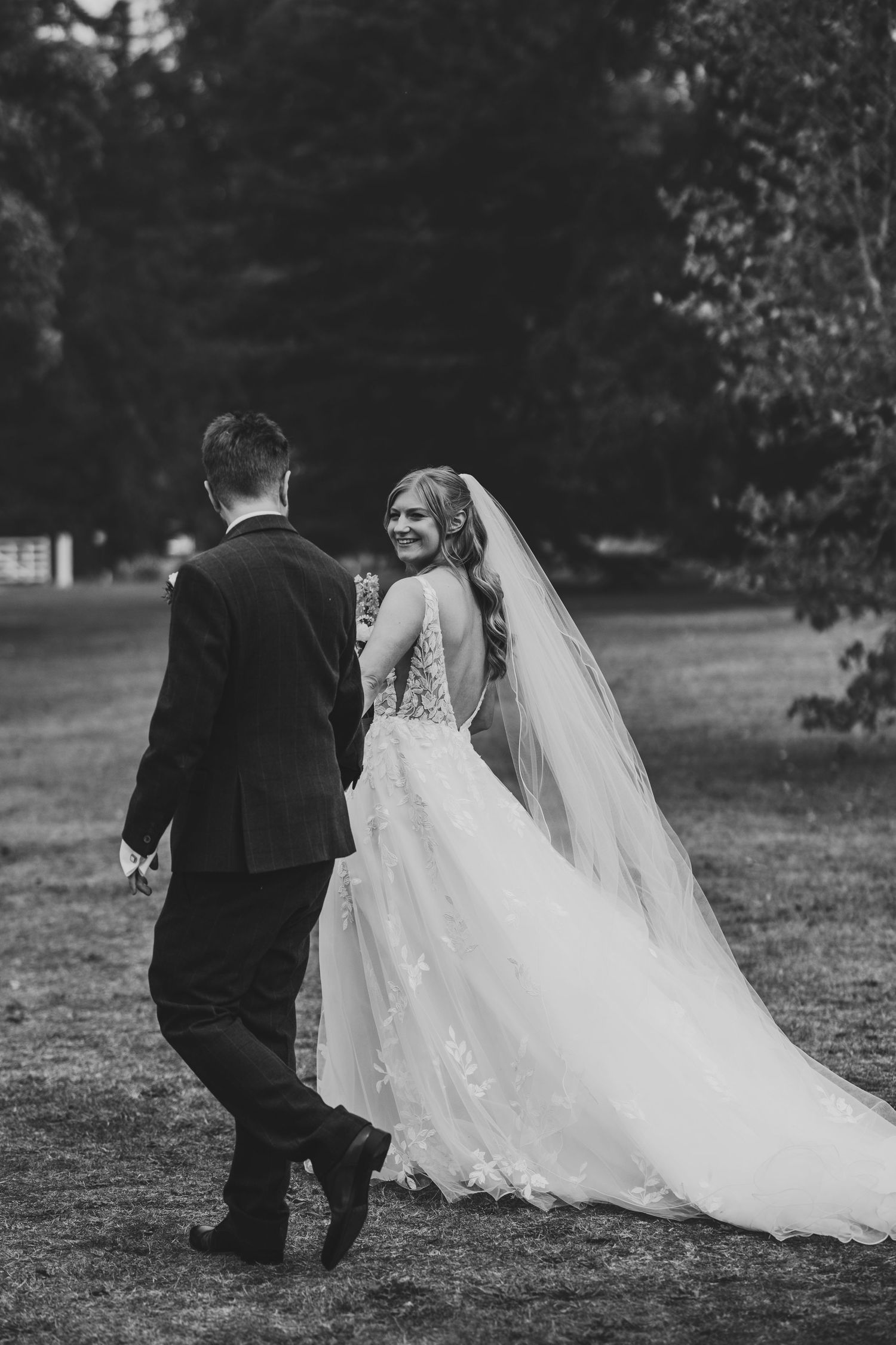 Black and white photo of a smiling bride in a lace gown and veil walking with a groom in a suit outdoors.